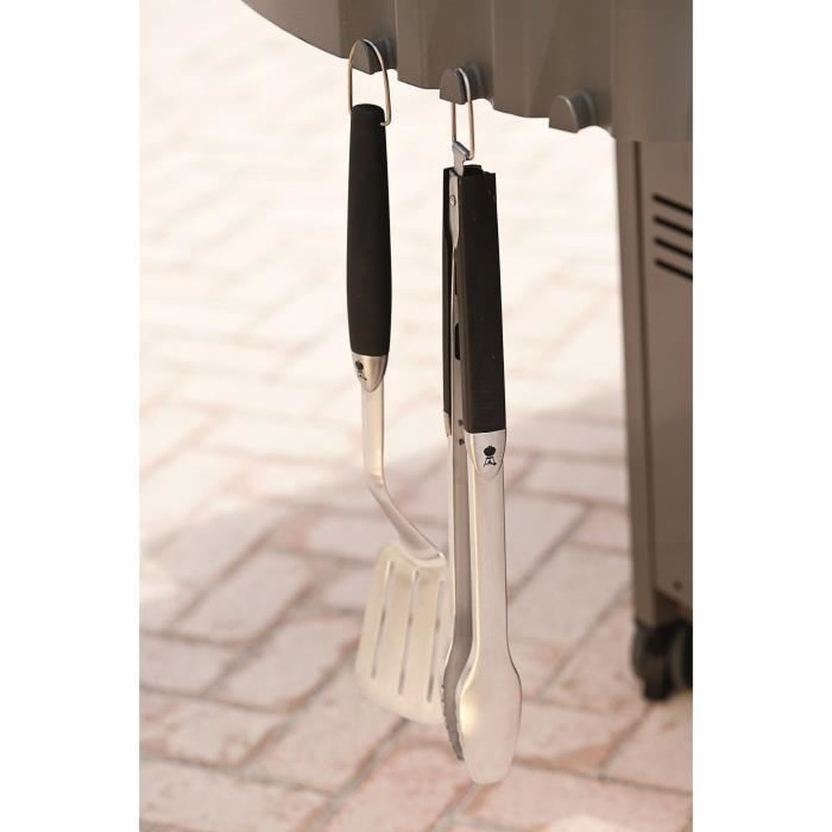 Narzędzia do grillowania wiszące na grillu: szpatułka i szczypce z czarnymi uchwytami.