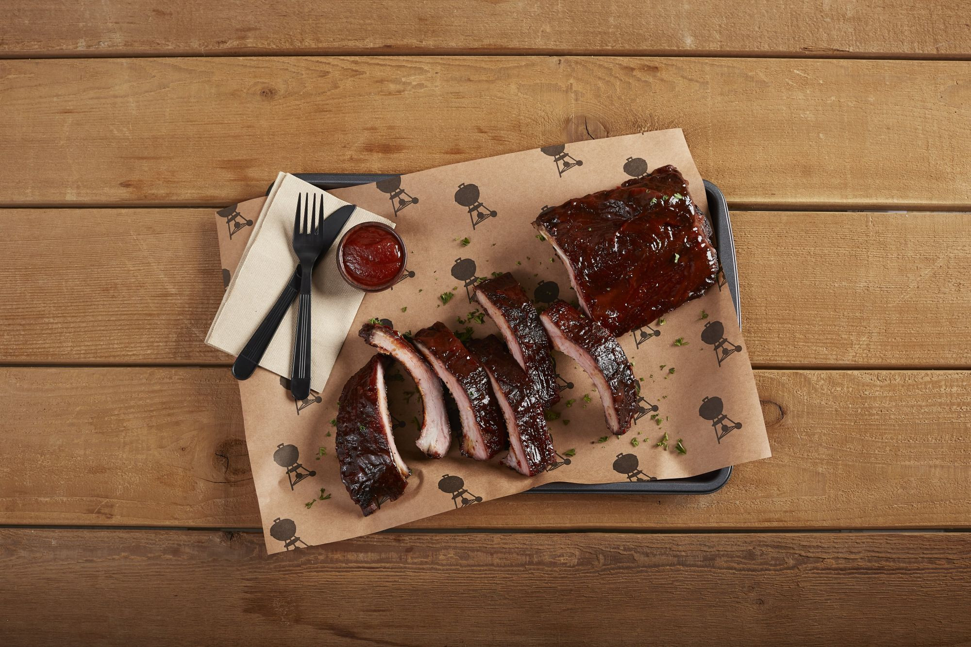 Costillas en rodajas en una bandeja con salsa barbacoa, tenedor, cuchillo y servilleta sobre una mesa de madera.