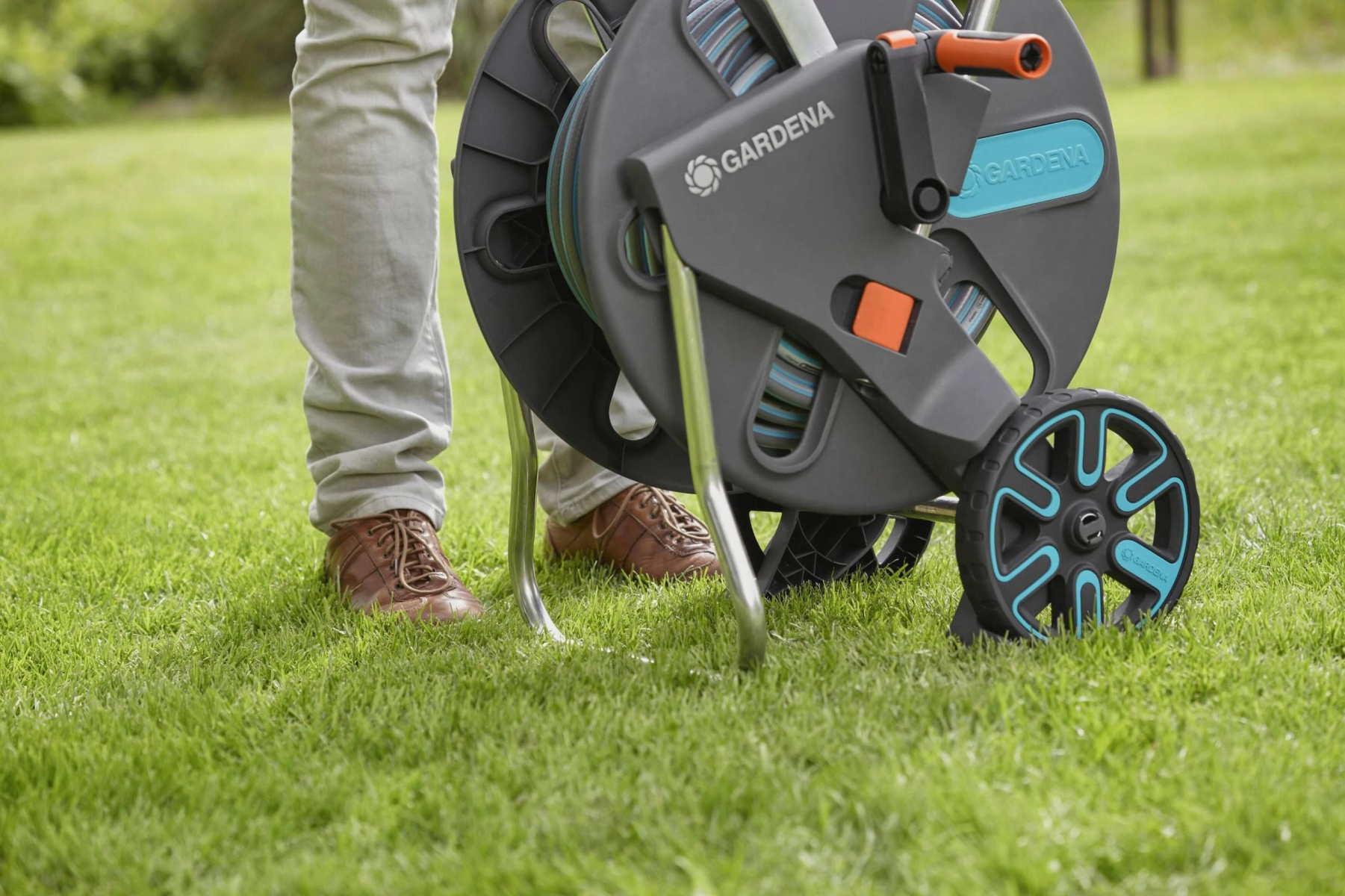 Mann, der eine Schlauchtrommel auf Gras rollt. Die Trommel ist schwarz und blaugrün, mit einem Griff und Rädern. Braune Schuhe sichtbar.