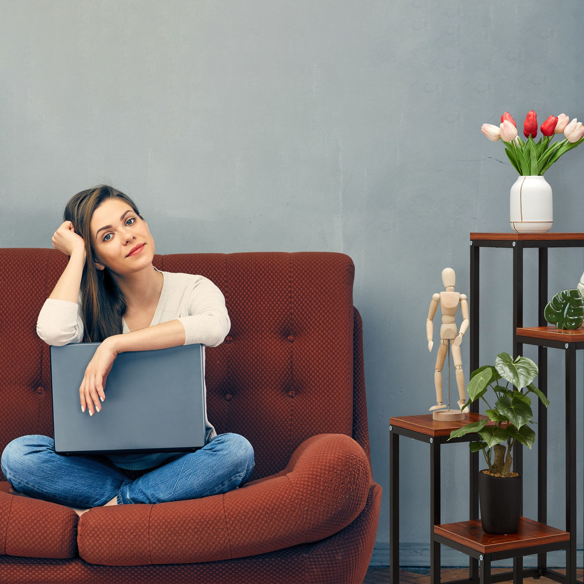 Frau auf rotem Sofa mit Laptop, gestaffelter Pflanzenständer mit Pflanzen und Holzfigur an grauer Wand.