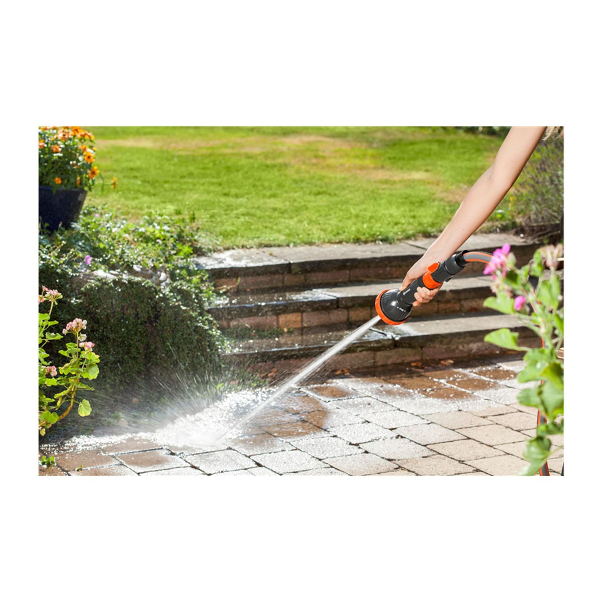 Una persona spruzza acqua su un patio in mattoni con un tubo, vicino a fiori ed erba.
