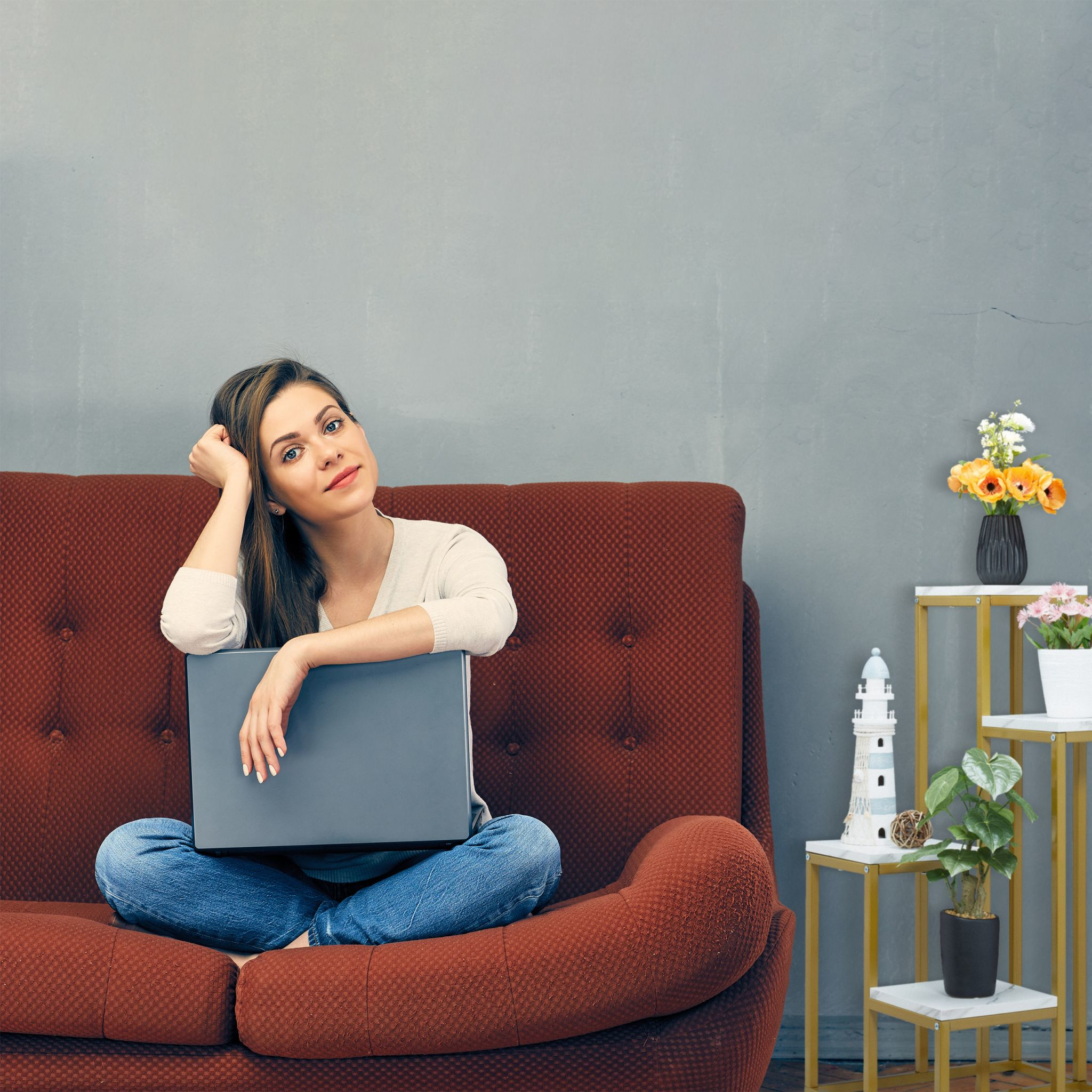 Frau auf einem Sofa mit Laptop, in der Nähe eines Pflanzenständers mit Blumen.