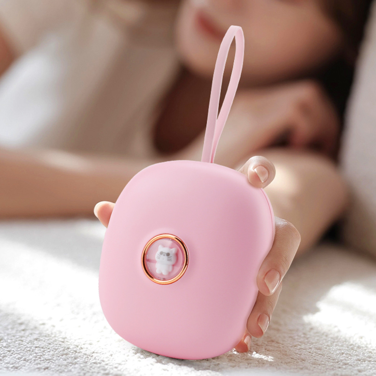 Een roze, ovale handwarmer met kattenontwerp, vastgehouden door een hand. Een persoon is wazig op de achtergrond.