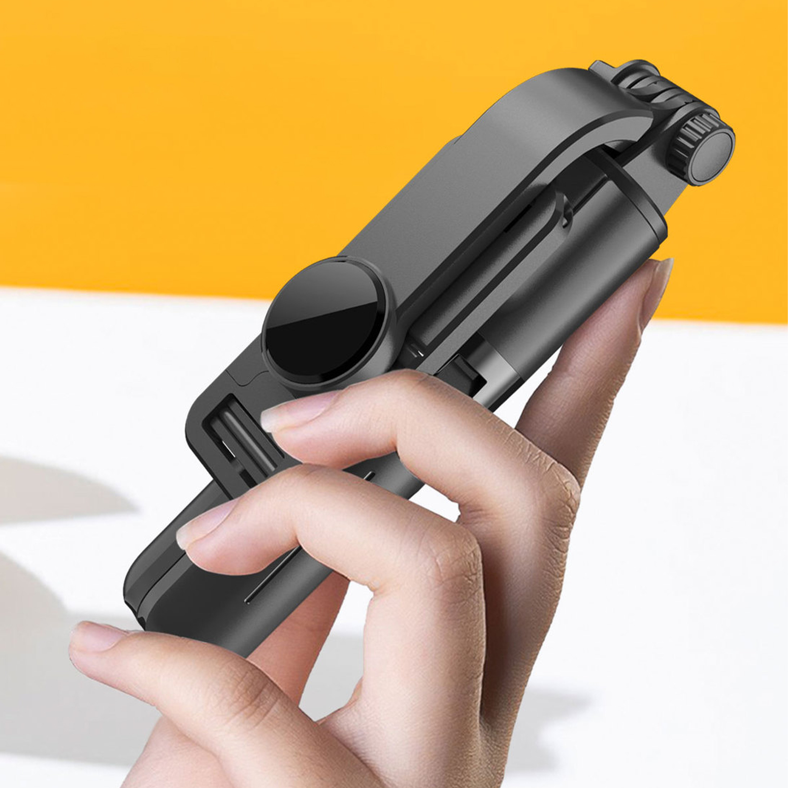 Hand hält schwarzen, faltbaren Selfie-Stick, gelber Hintergrund.
