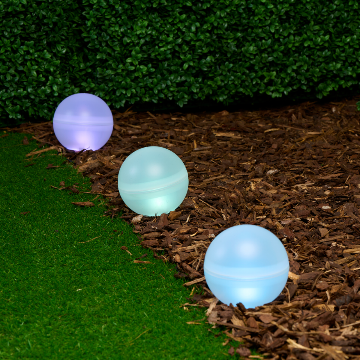 Three illuminated orbs on a bed of mulch. Green grass and hedge in background.