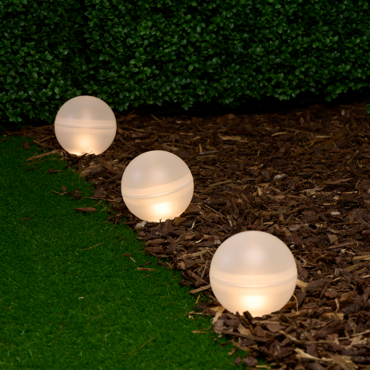Three illuminated white spheres are on mulch, in front of a green hedge, creating an appealing scene.