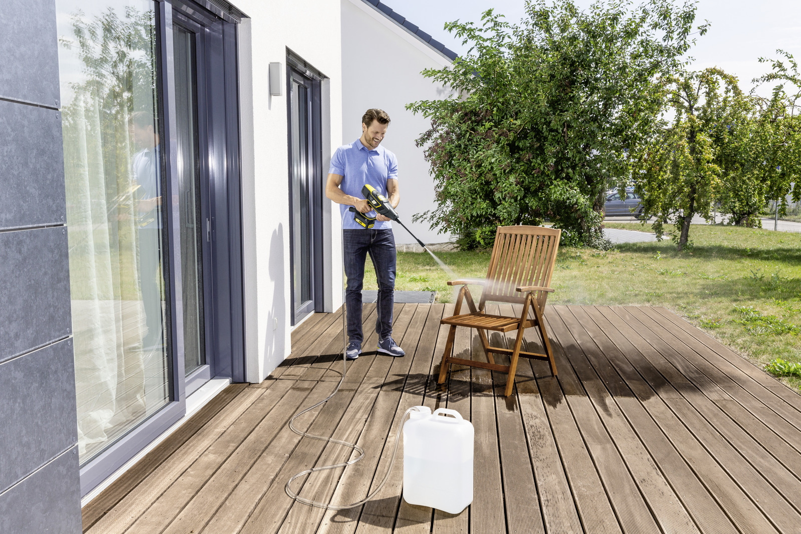 Un hombre usa una hidrolimpiadora en una silla de madera en una terraza. Un recipiente de agua está cerca.