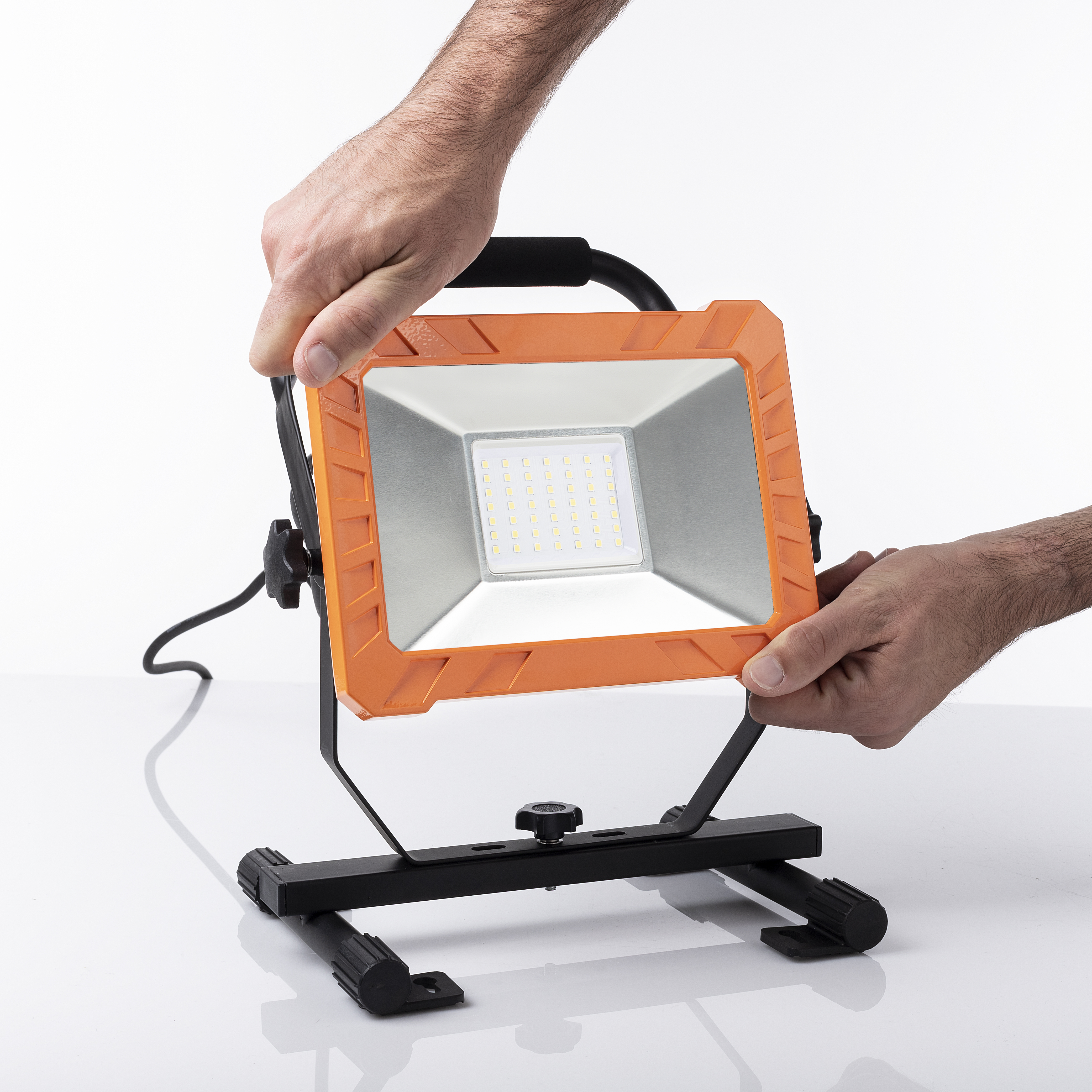 Twee handen houden de oranje en zwarte werklamp vast. Het licht staat op een wit oppervlak.