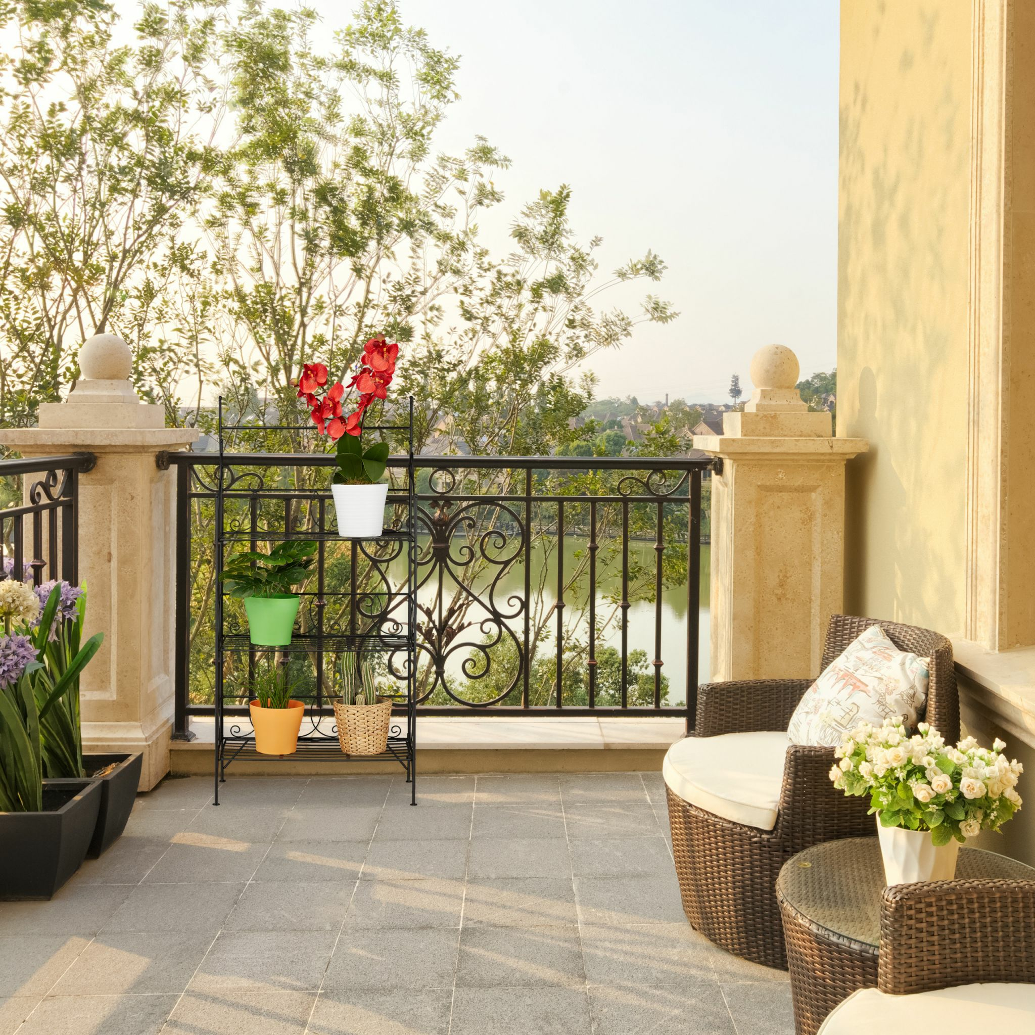 Balkon mit schwarzem Geländer, Pflanzenständer mit Blumen und Korbmöbeln, Blick auf Wasser und Bäume.