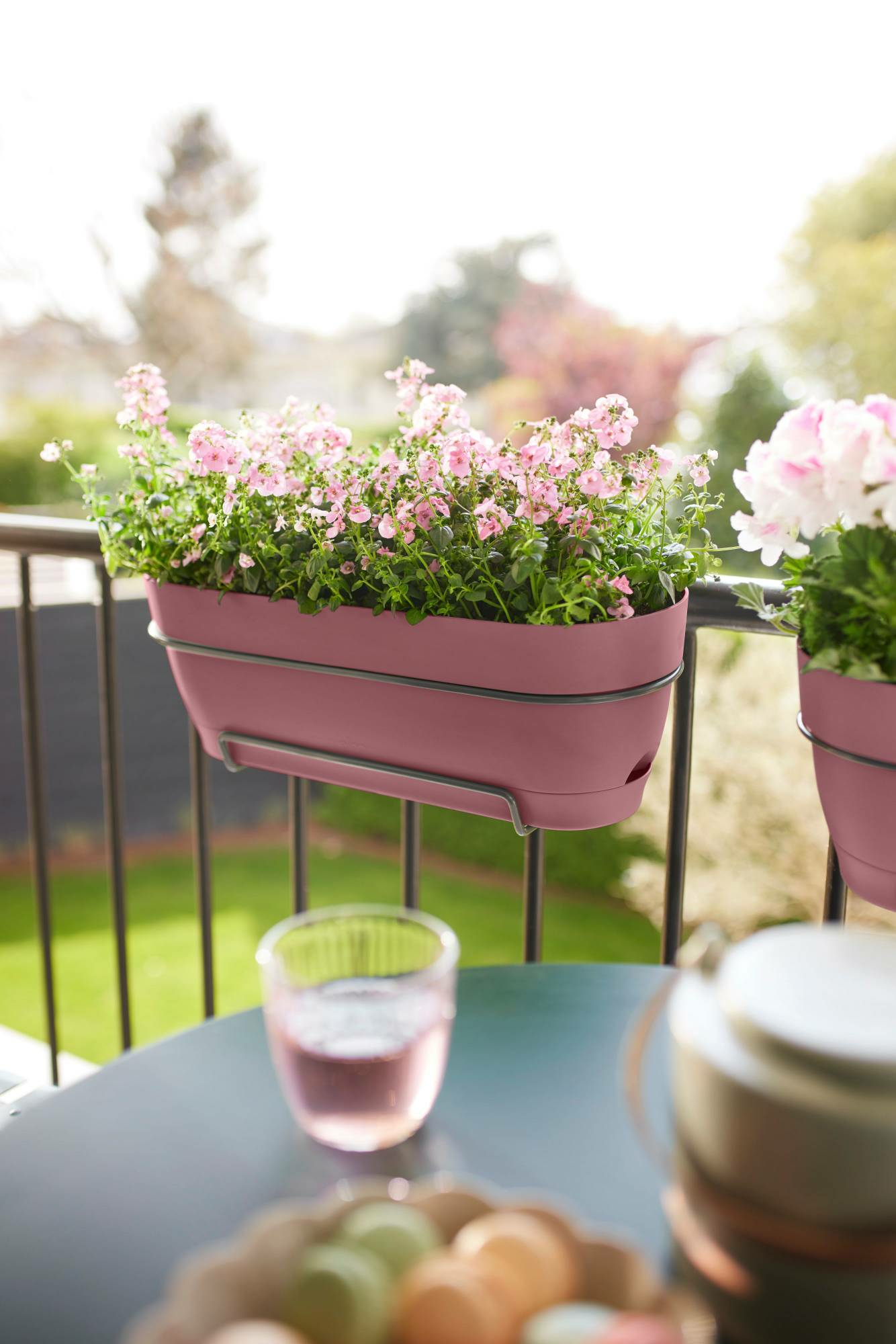 Scena z balkonu z różową donicą, kwiatami i szklanką różowego płynu na stole.