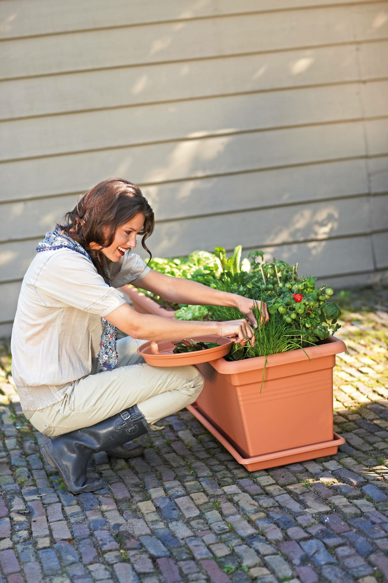 Una mujer cuidando plantas en un jardín. Sostiene una bandeja y sonríe.