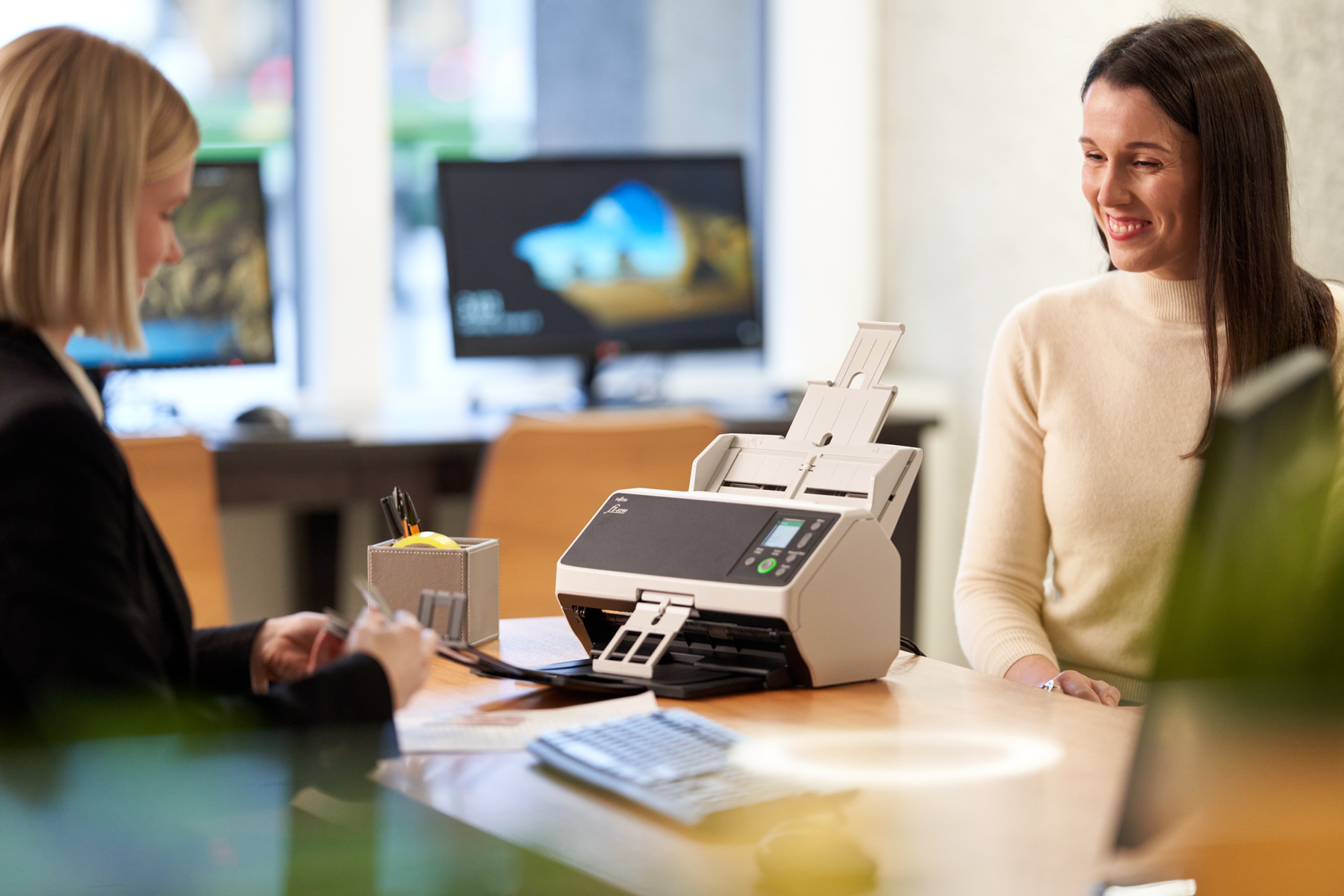 Dwie kobiety w biurze. Jedna skanuje dokumenty za pomocą beżowego skanera Fujitsu na biurku.