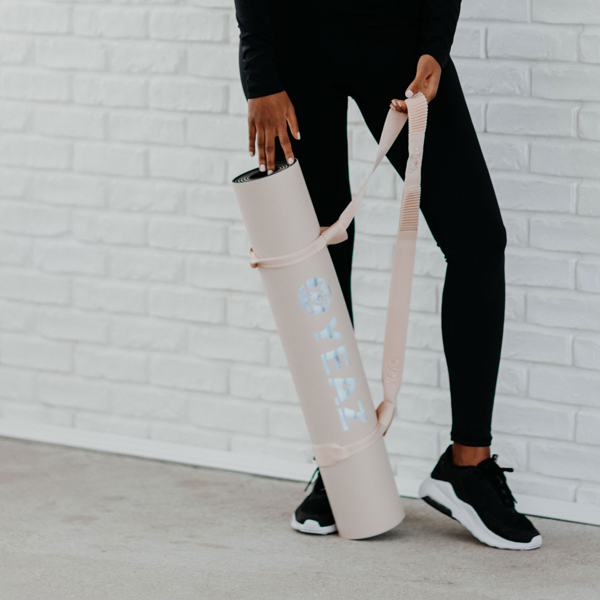 Person hält eine gerollte Yogamatte mit einem Gurt, trägt schwarze Leggings und Schuhe, steht an einer Wand.