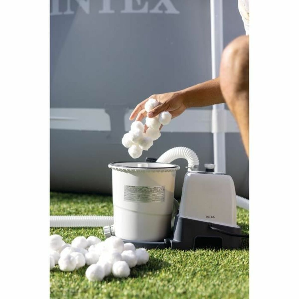 Persoon voegt witte filterballen toe aan een witte container naast een zwembadfiltersysteem op groen gras.