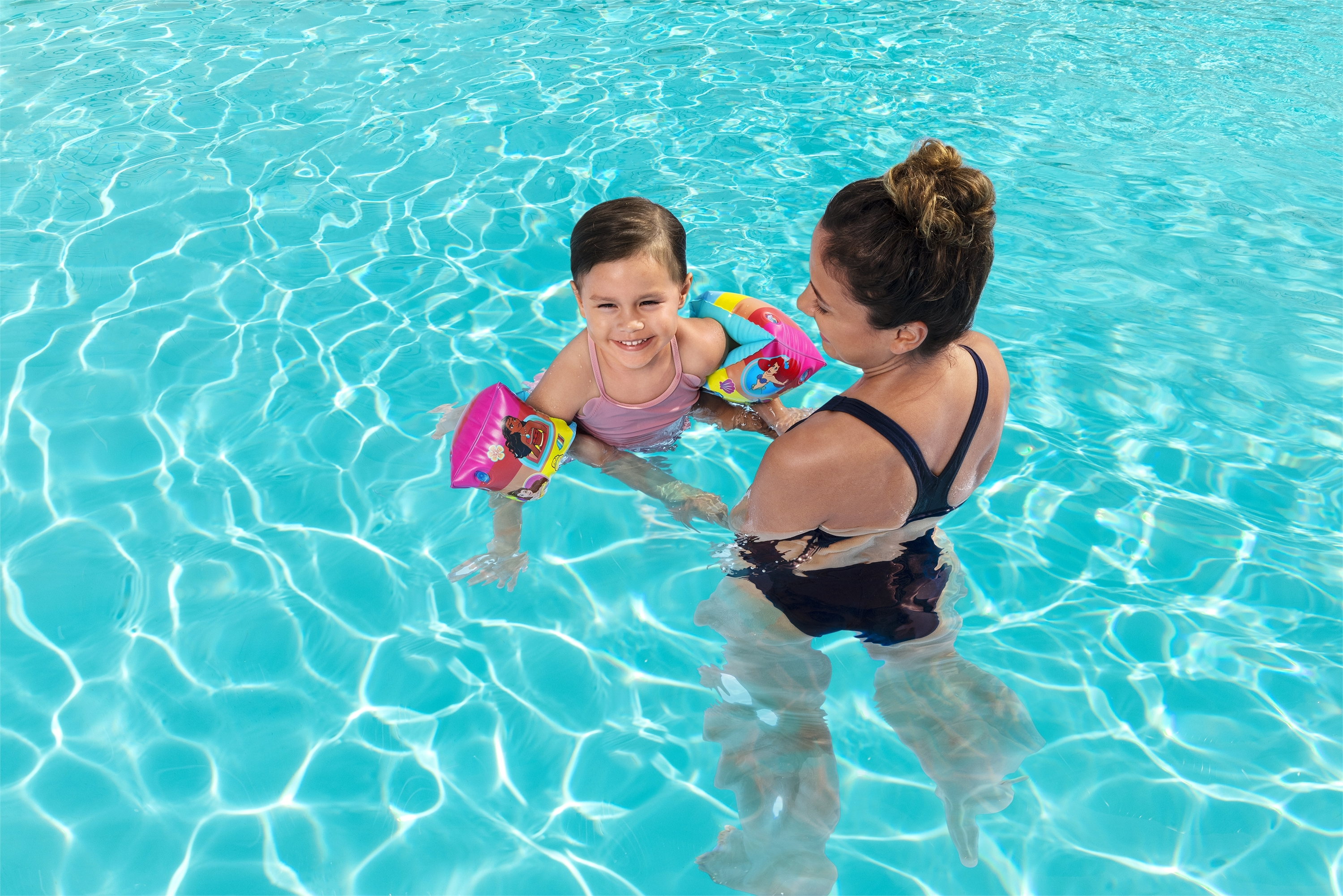 Mujer en piscina sujetando a niña con flotadores, ambas sonriendo, agua azul clara.
