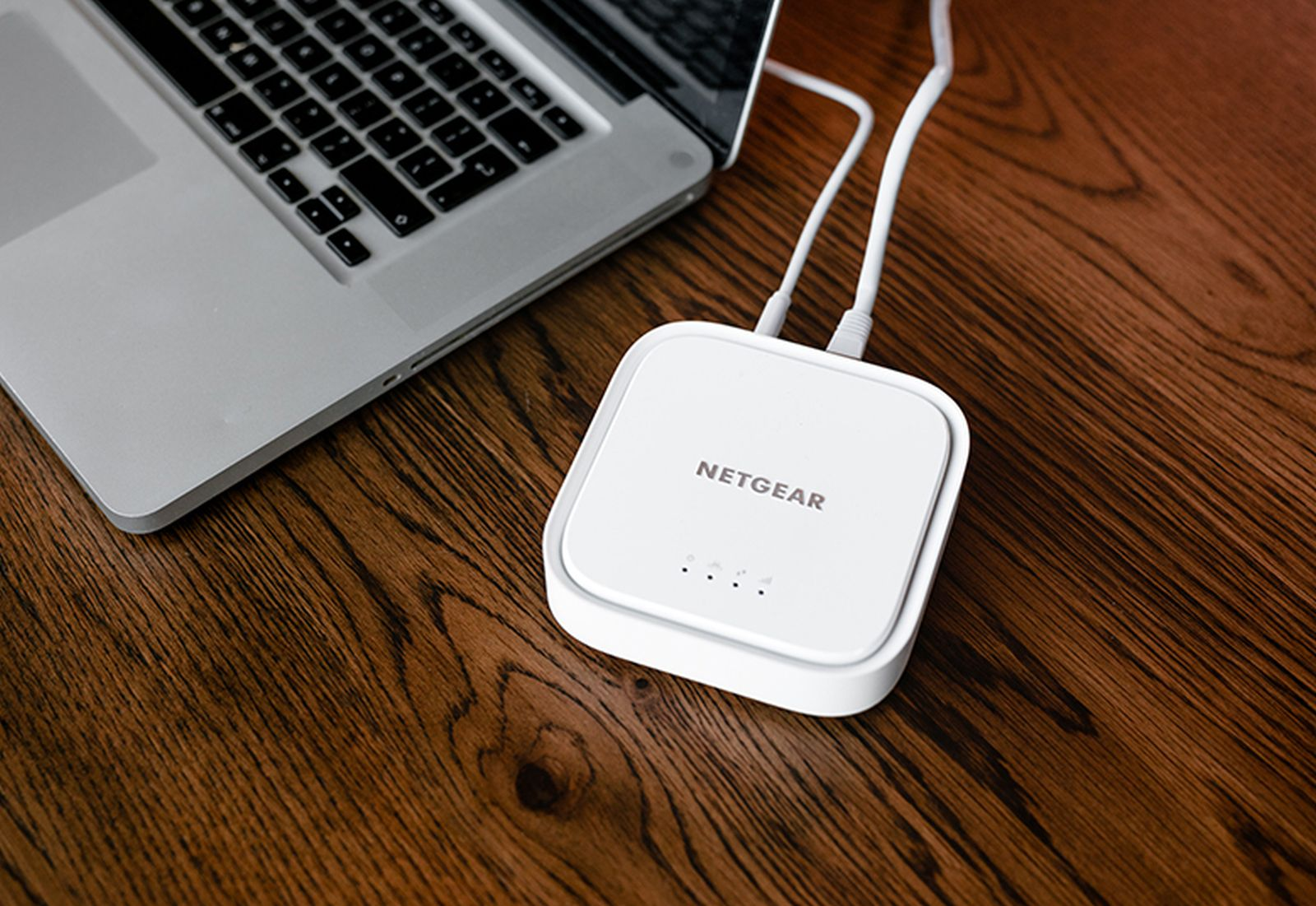 Un enrutador Netgear blanco se encuentra sobre una mesa de madera junto a una laptop, conectado con cables.