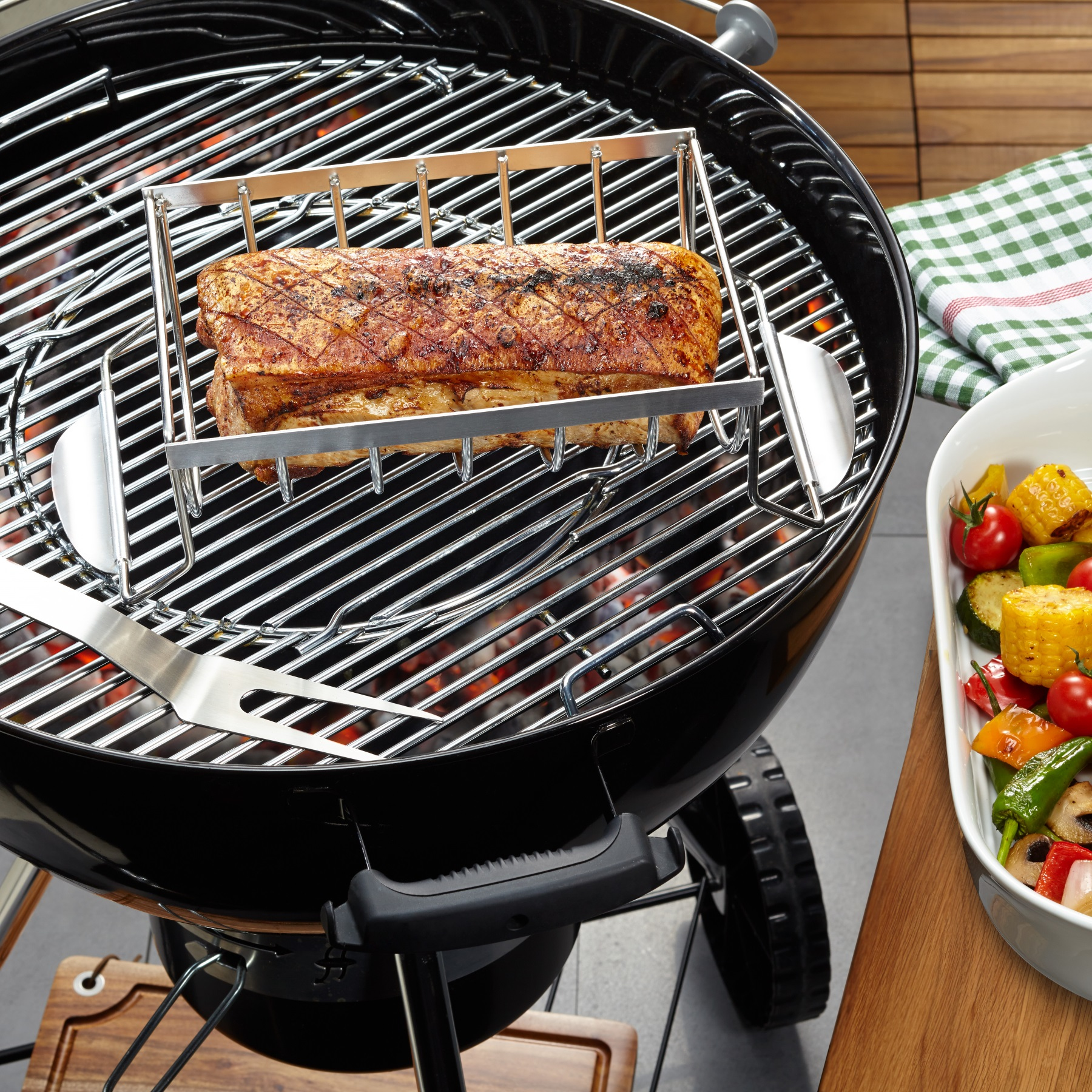 Ein Grill mit einem großen Fleischstück, einem Rost und Gemüse in einer weißen Schüssel.