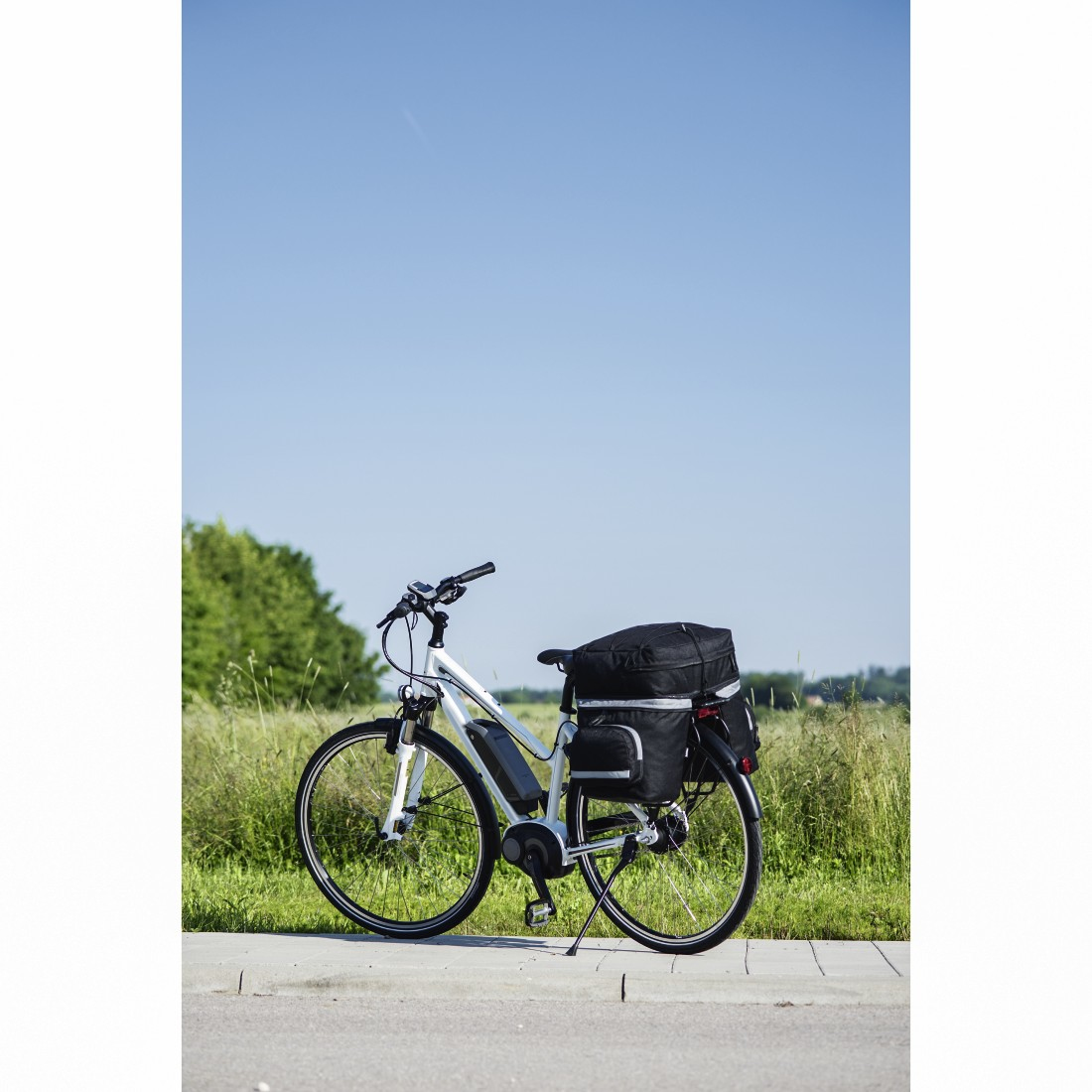 Weißes Fahrrad mit schwarzem Gepäck hinten, geparkt neben einer Straße mit blauem Himmel.