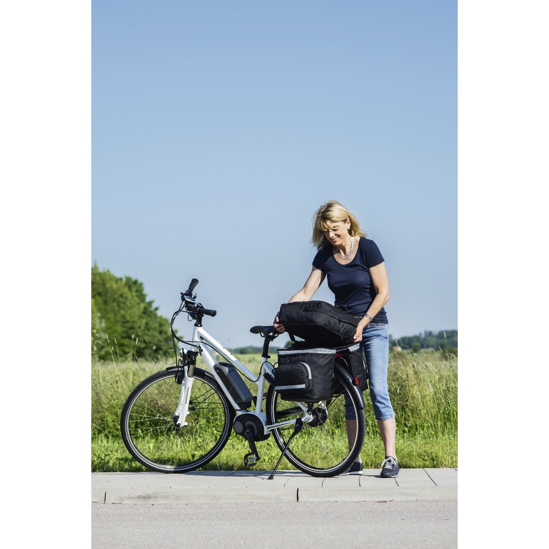 Eine Frau mit Fahrrad, packt eine Tasche aus. Fahrrad weiß, Taschen schwarz, blauer Himmel.