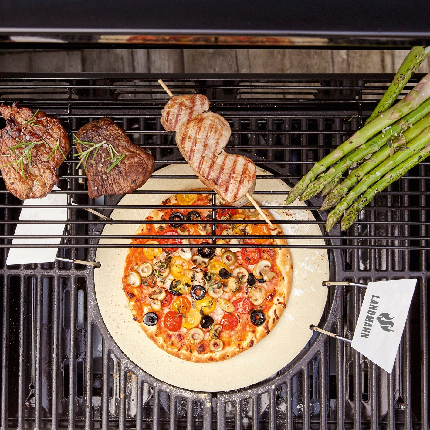 Grill mit Steak, Spießen, Spargel und Pizza. Markenlogos sind sichtbar.