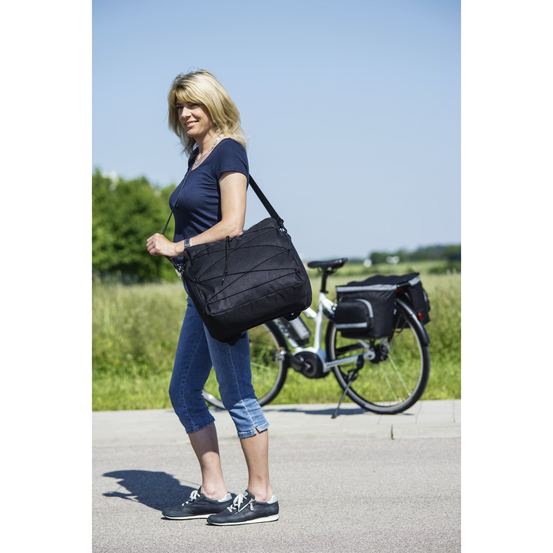Eine Frau geht mit einer schwarzen Tasche über der Schulter, im Hintergrund ein Fahrrad, sonniger Tag.