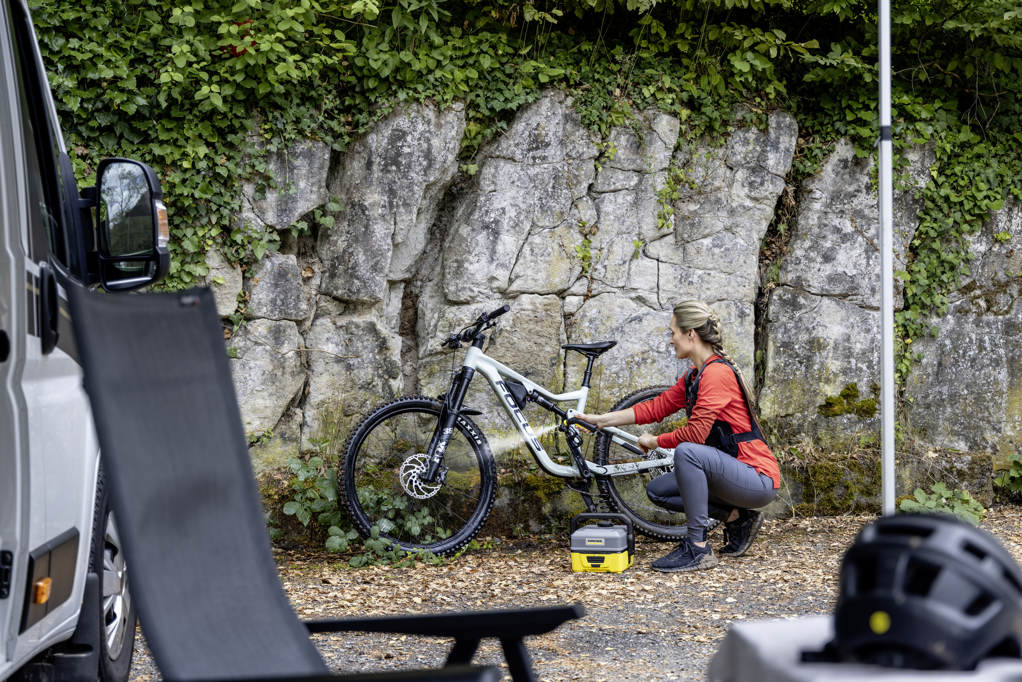 Une femme en chemise rouge et pantalon gris travaille sur un vélo. On voit un van, des arbres et des rochers.
