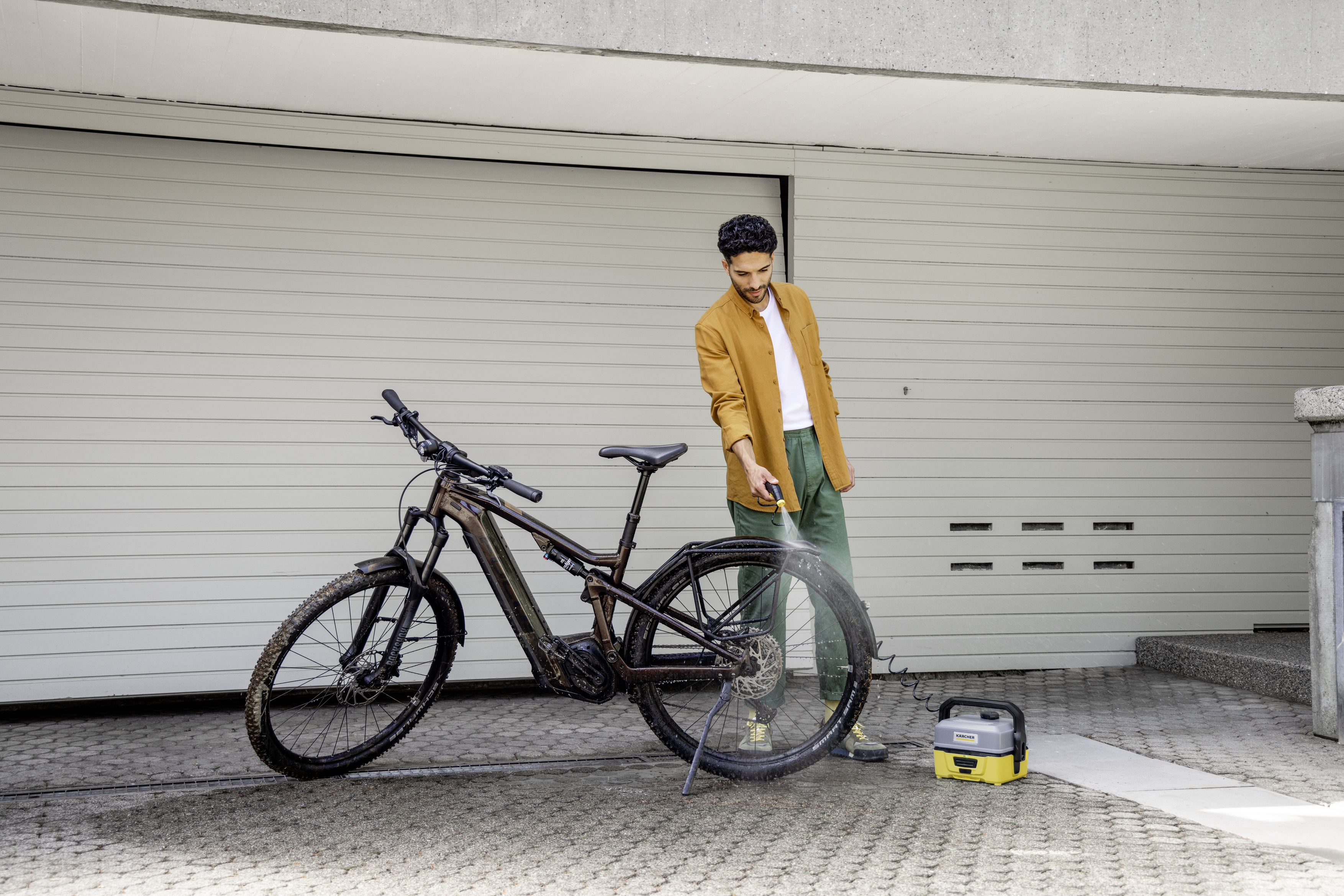 Homme lavant un vélo avec un nettoyeur haute pression. Le vélo est marron. La porte de garage est le fond.