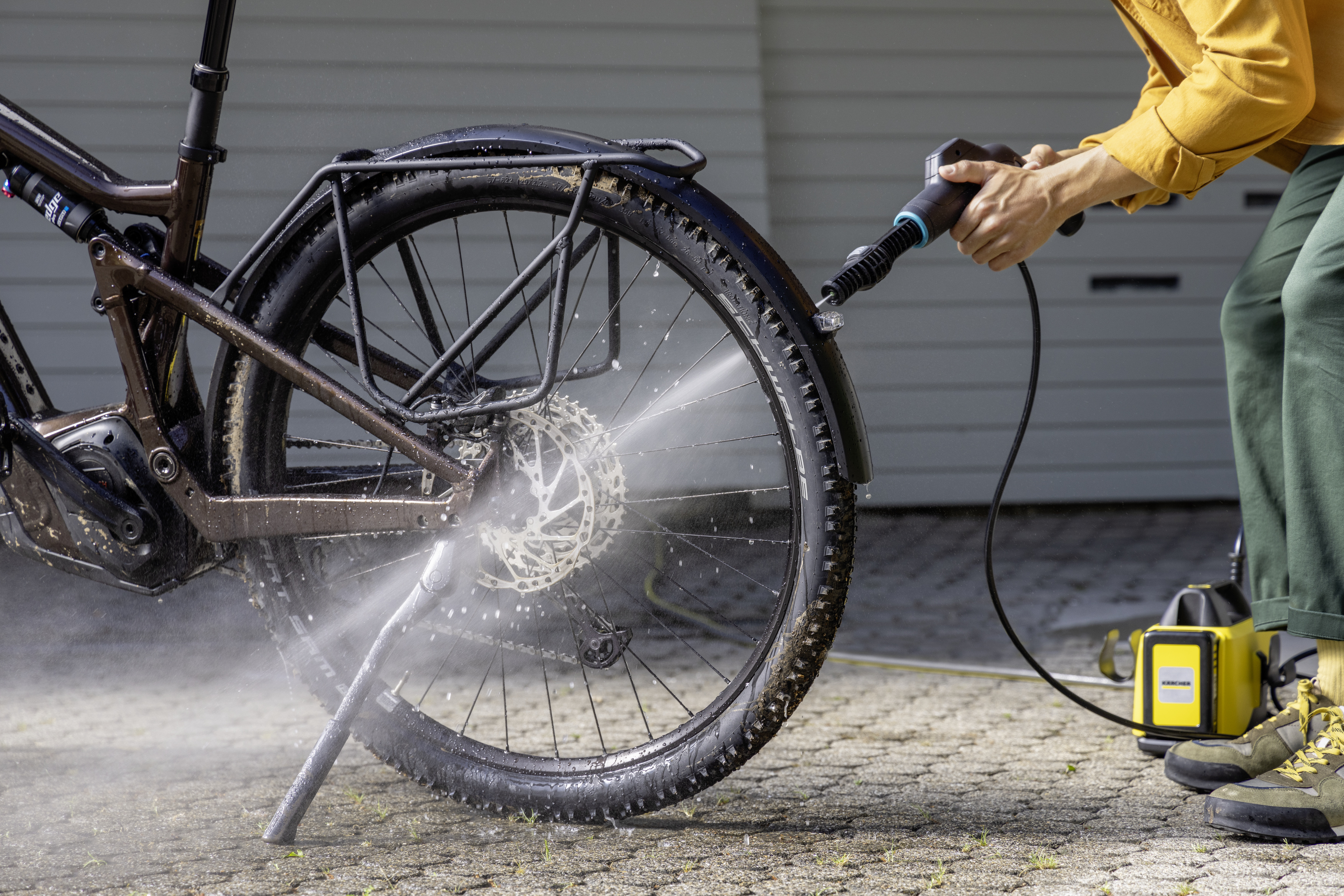 Person washing a bicycle wheel with a pressure washer. The bike is brown, and the setting is outdoors.