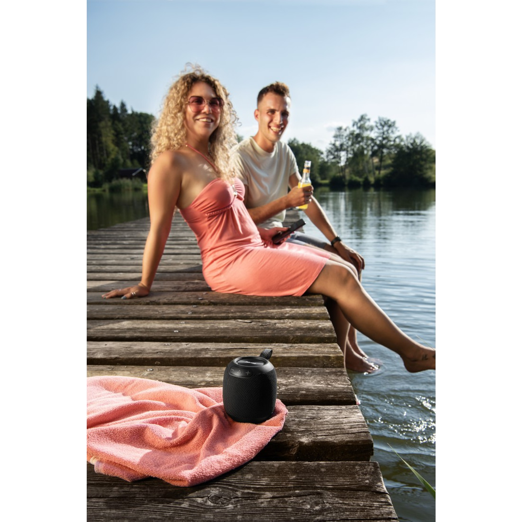 Dwie osoby siedzące na pomoście, kobieta w różowej sukience i mężczyzna, z głośnikiem i widokiem na jezioro.