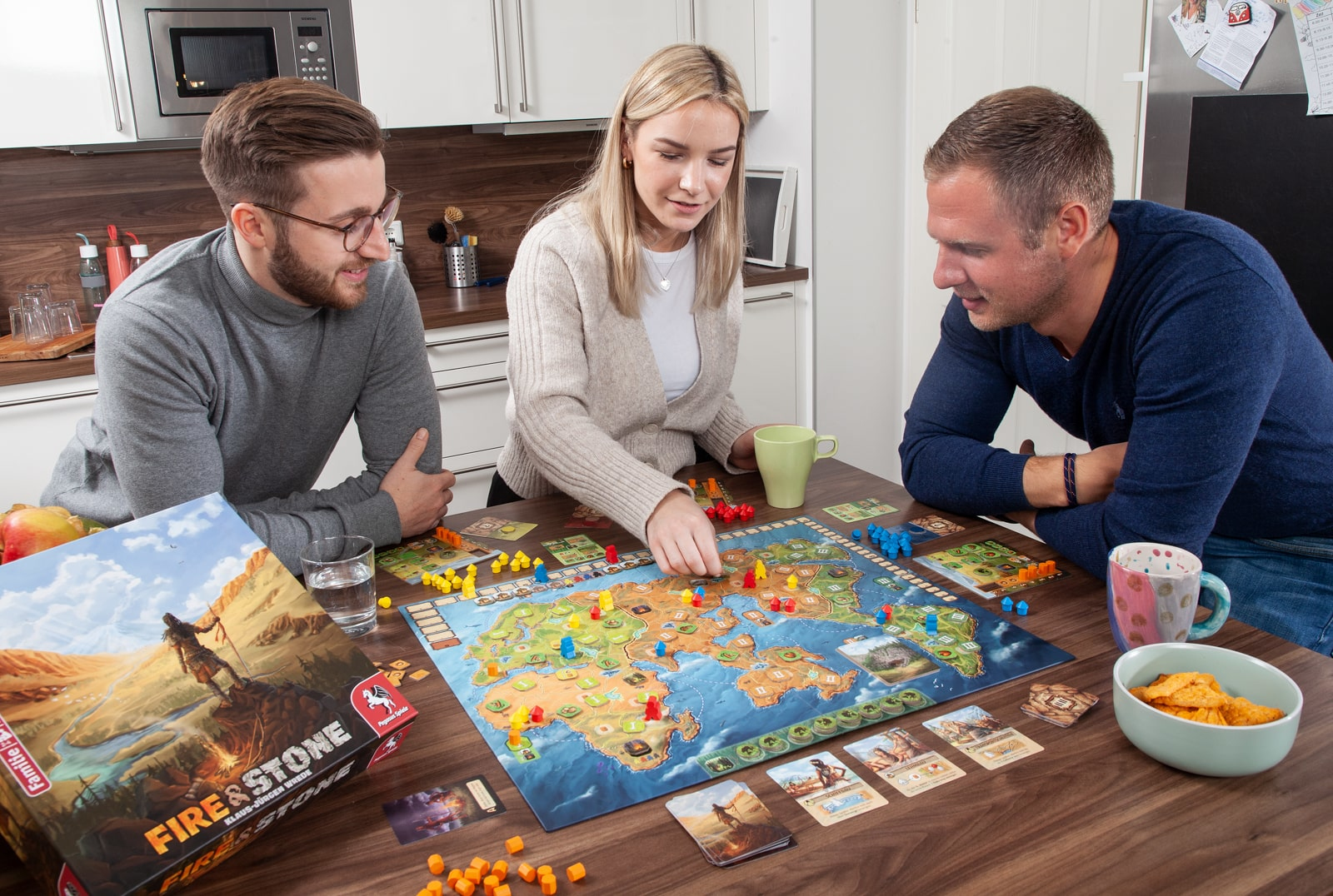 Drei Personen spielen ein Brettspiel. Das Spielbrett ist auf einem Holztisch in der Küche.