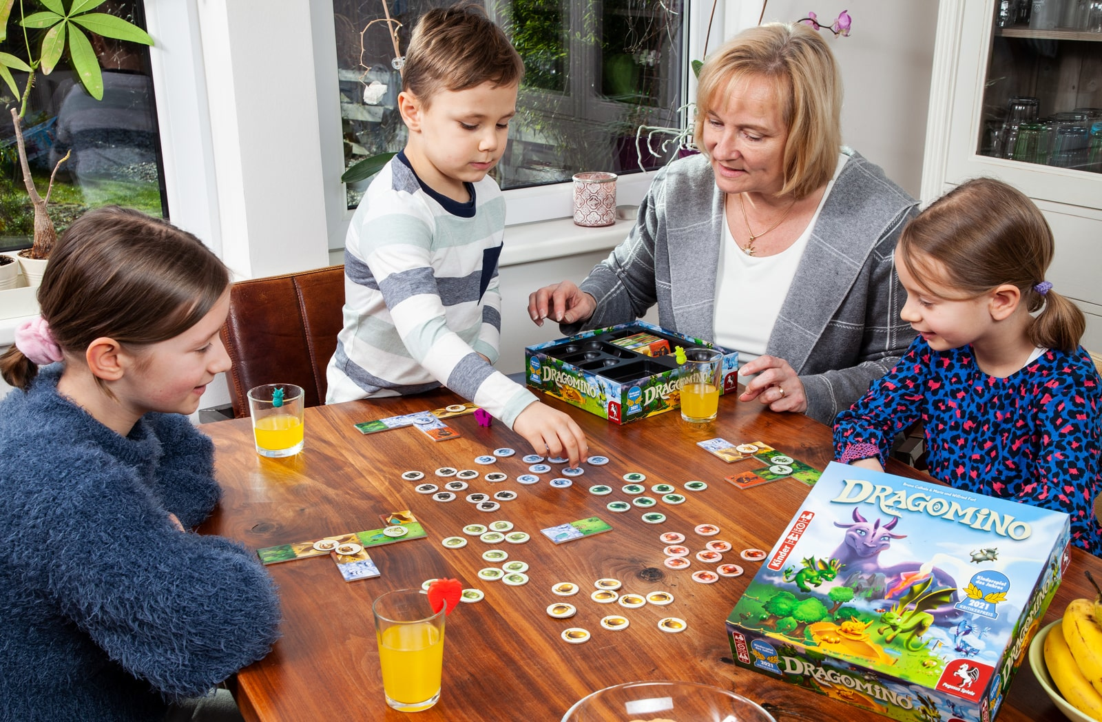 Cztery osoby grające w grę planszową przy drewnianym stole. Szklanki z sokiem i banan.