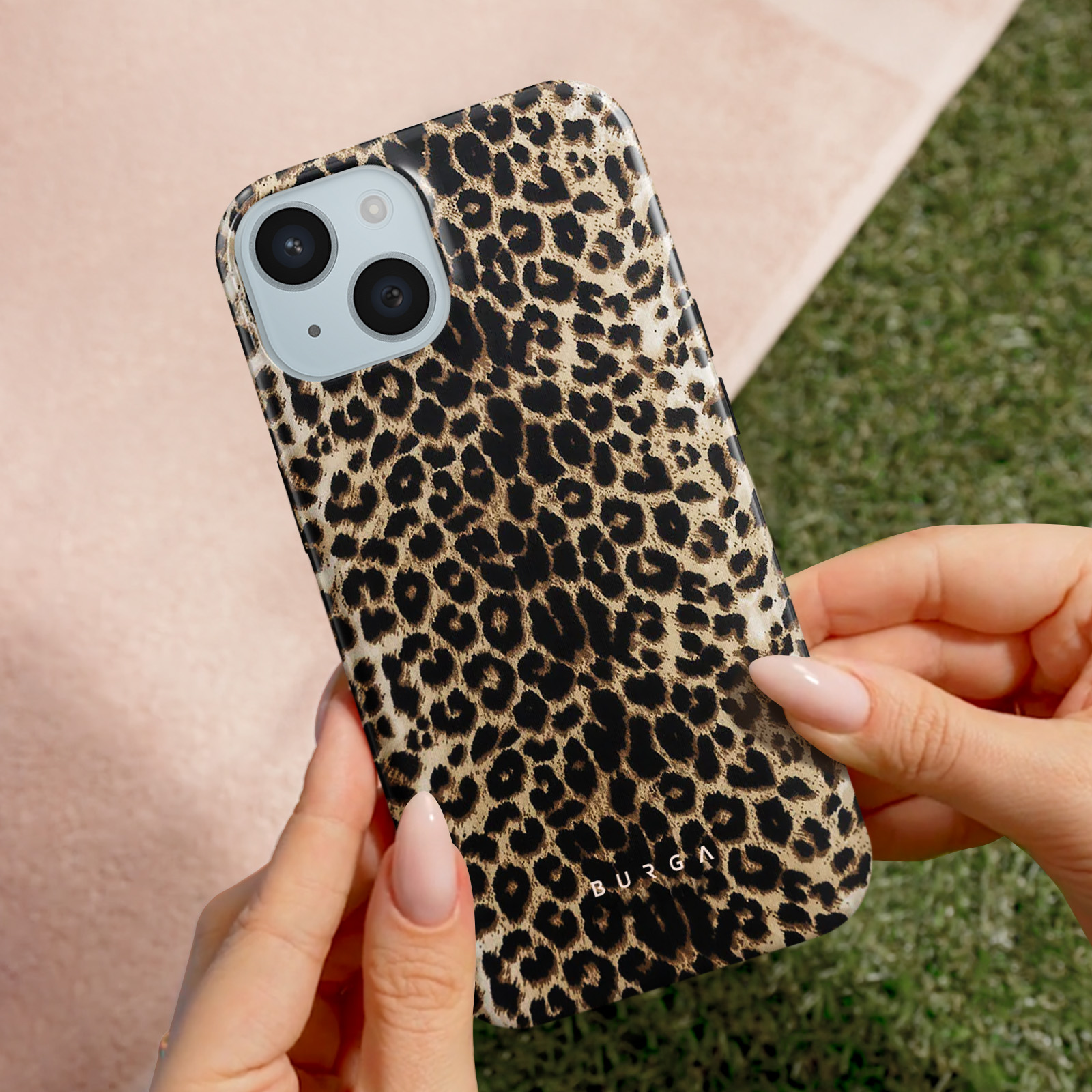 Een telefoon met een luipaardprint hoes wordt vastgehouden door handen. Achtergrond is groen gras en roze stof.