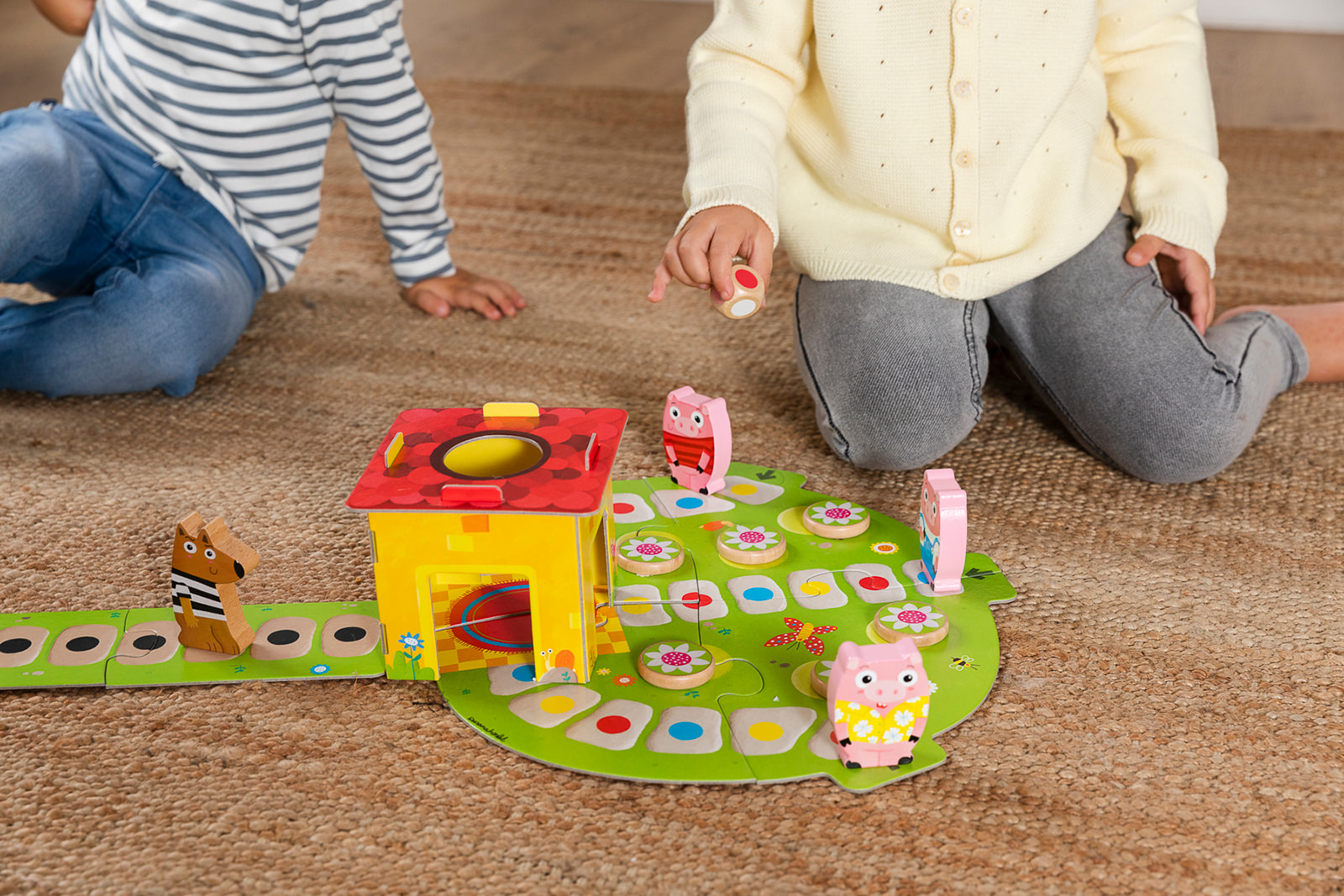 Dwoje dzieci grających w grę planszową z drewnianymi figurkami. Domek i kwiaty na zielonej macie.