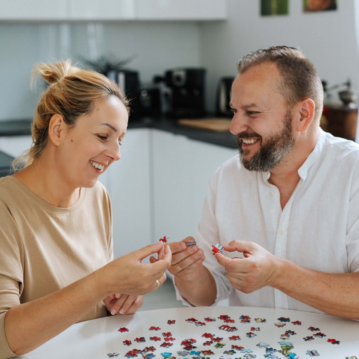 Dwie uśmiechnięte osoby, kobieta i mężczyzna, układają puzzle przy białym stole.
