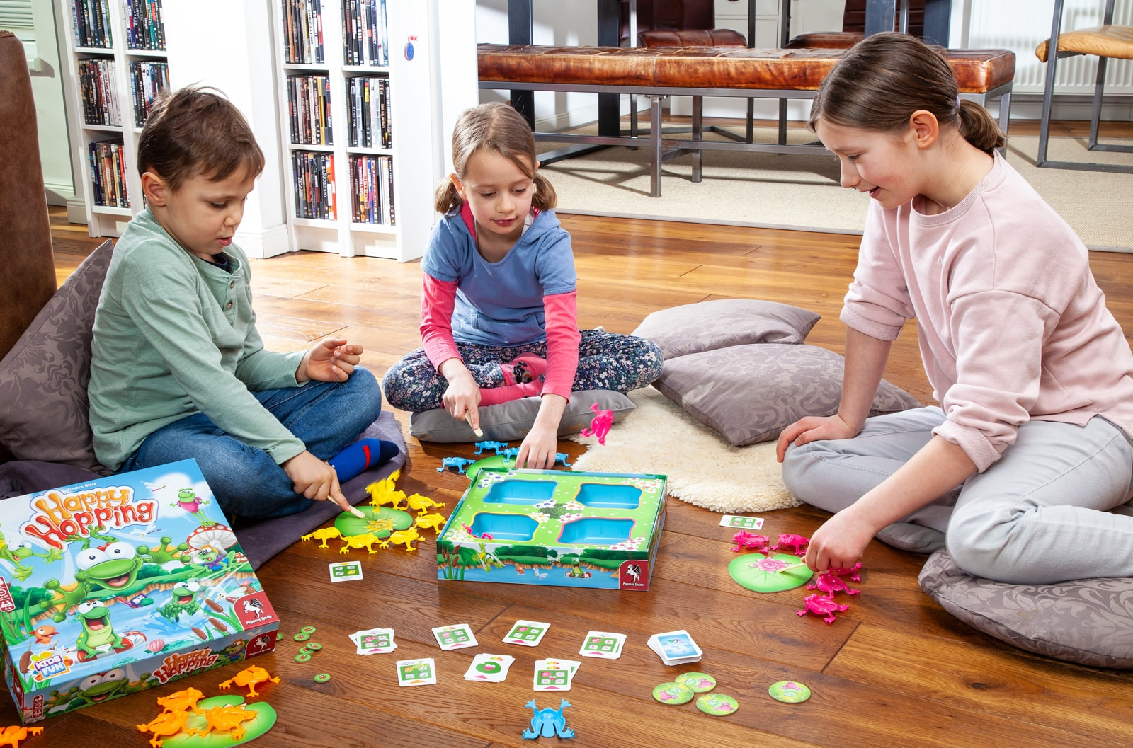 Drei Kinder spielen ein Brettspiel mit Fröschen. Das Spiel findet auf einem Holzboden statt.