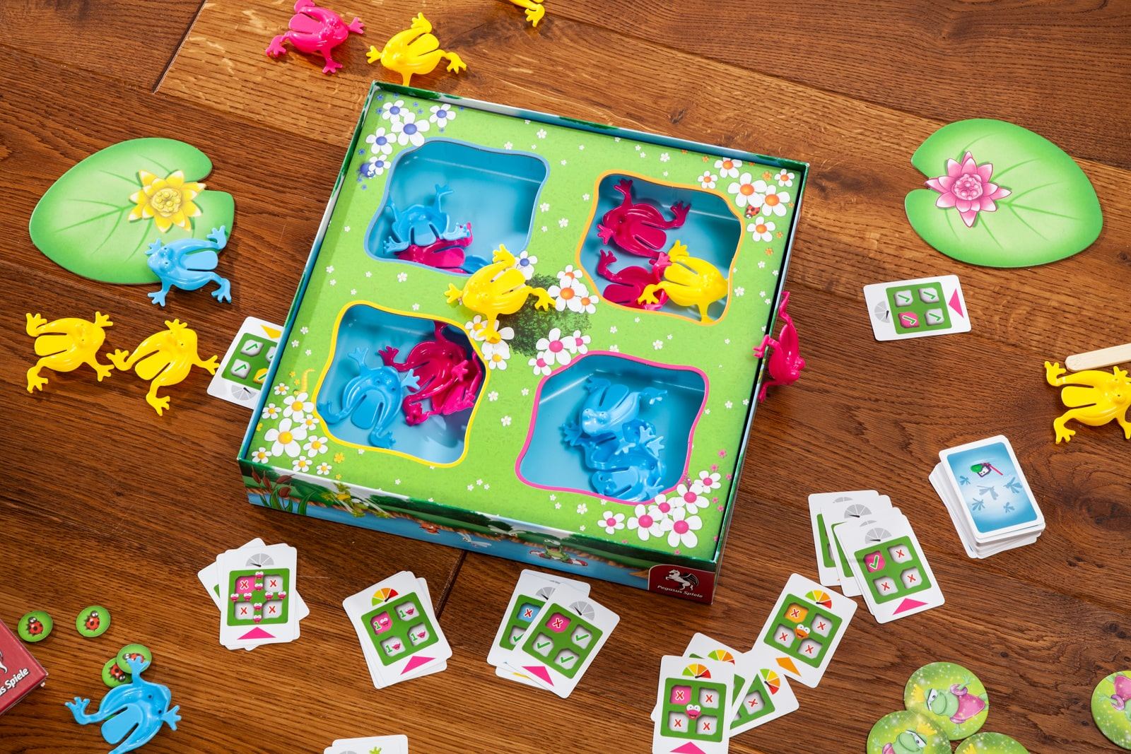 Brettspiel mit Fröschen auf Seerosenblättern. Schachtel und Spielsteine liegen auf Holz.