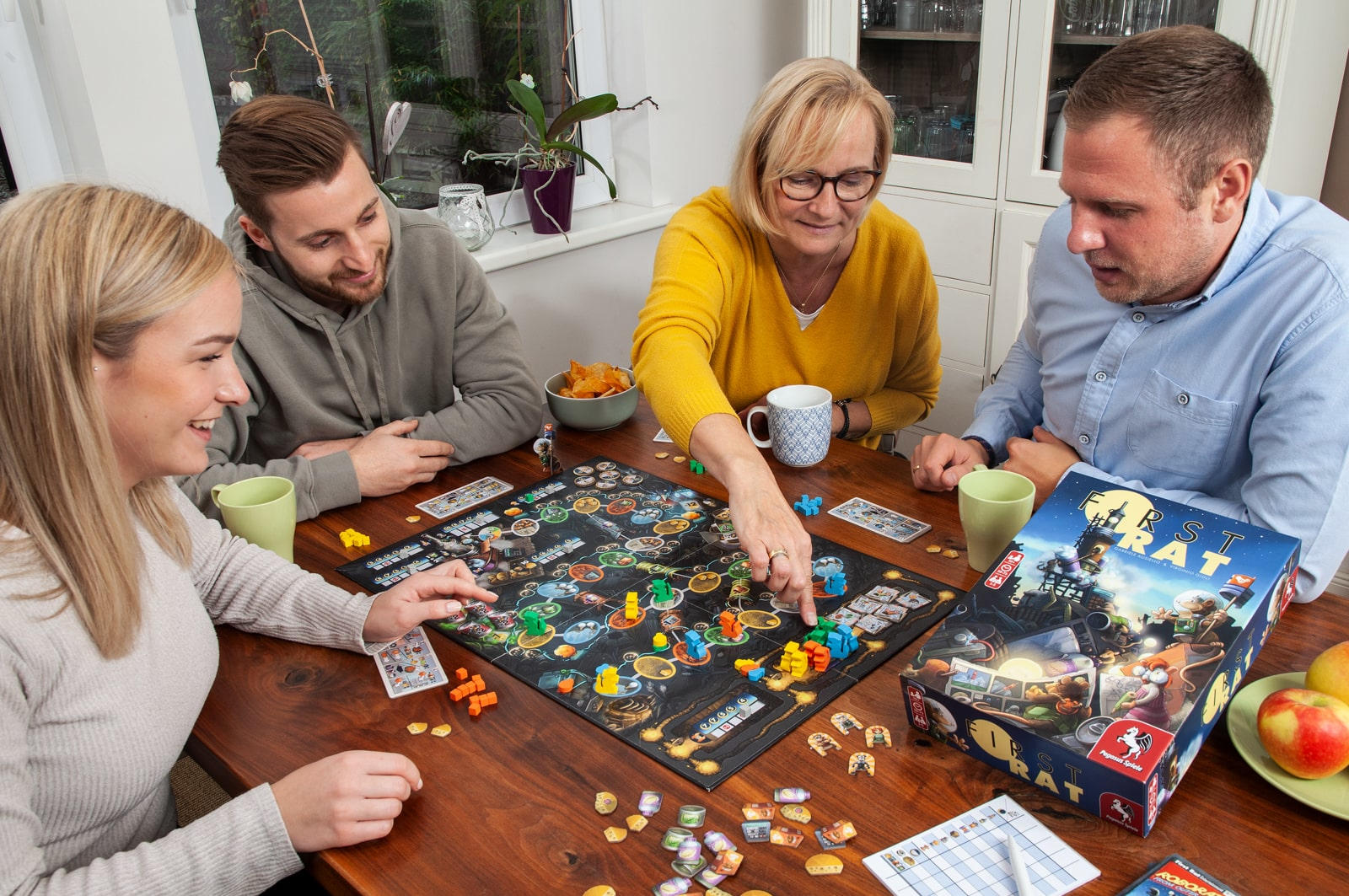 Vier Personen spielen ein Brettspiel. Das Spiel steht auf einem Holztisch. Äpfel und Tassen sind zu sehen.