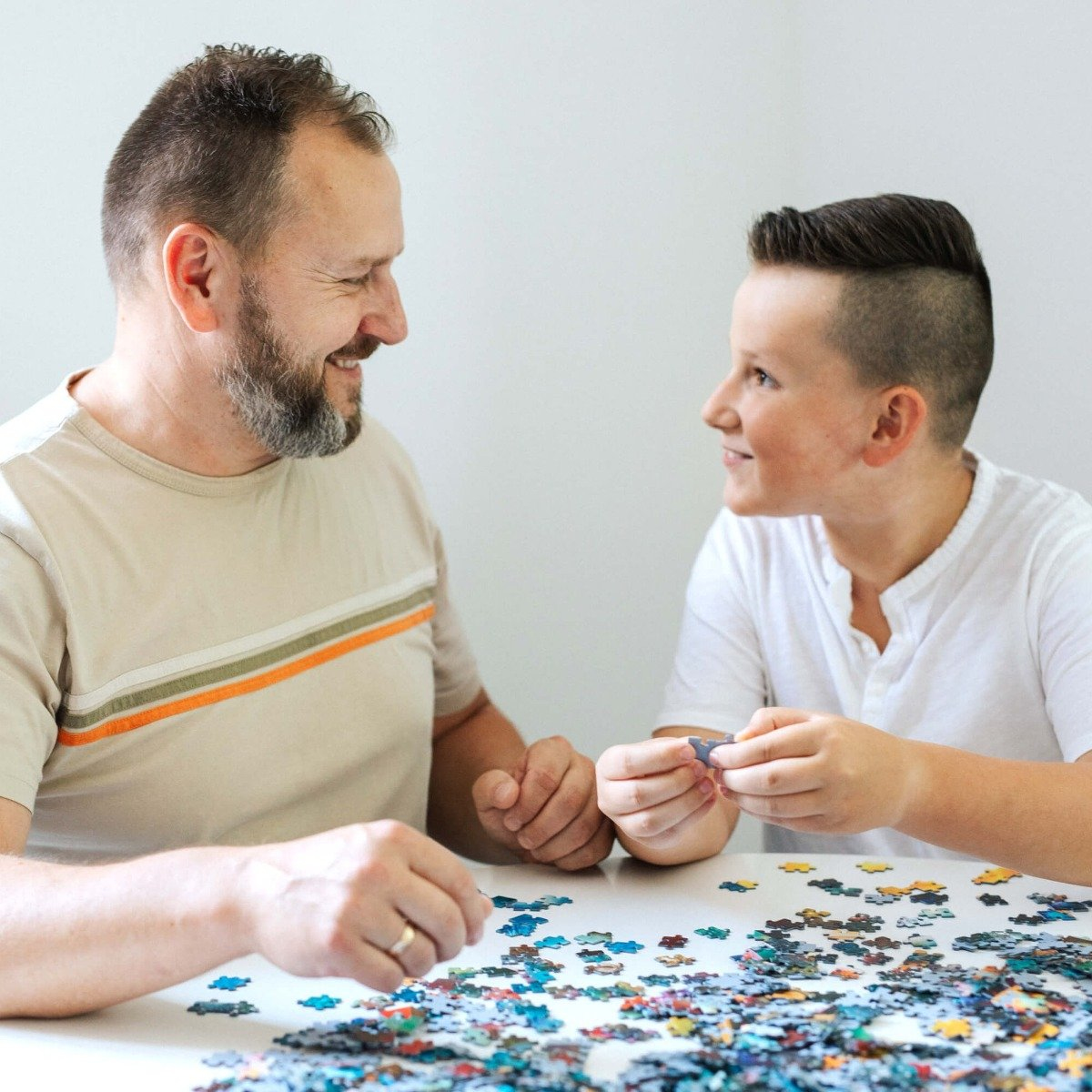 Un padre e un figlio stanno lavorando insieme a un puzzle su un tavolo bianco, sorridendo.