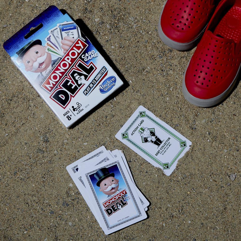 Pudełko do gry w karty Monopoly Deal i karty na piasku, obok czerwonych butów.