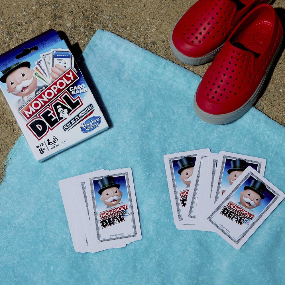 Gra karciana Monopoly Deal z kartami i czerwonymi butami na niebieskim ręczniku. Tło: piaszczysta powierzchnia.