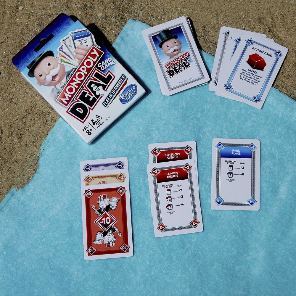 Gra karciana Monopoly Deal na niebieskim ręczniku i piasku. Karty rozłożone, pudełko widoczne.