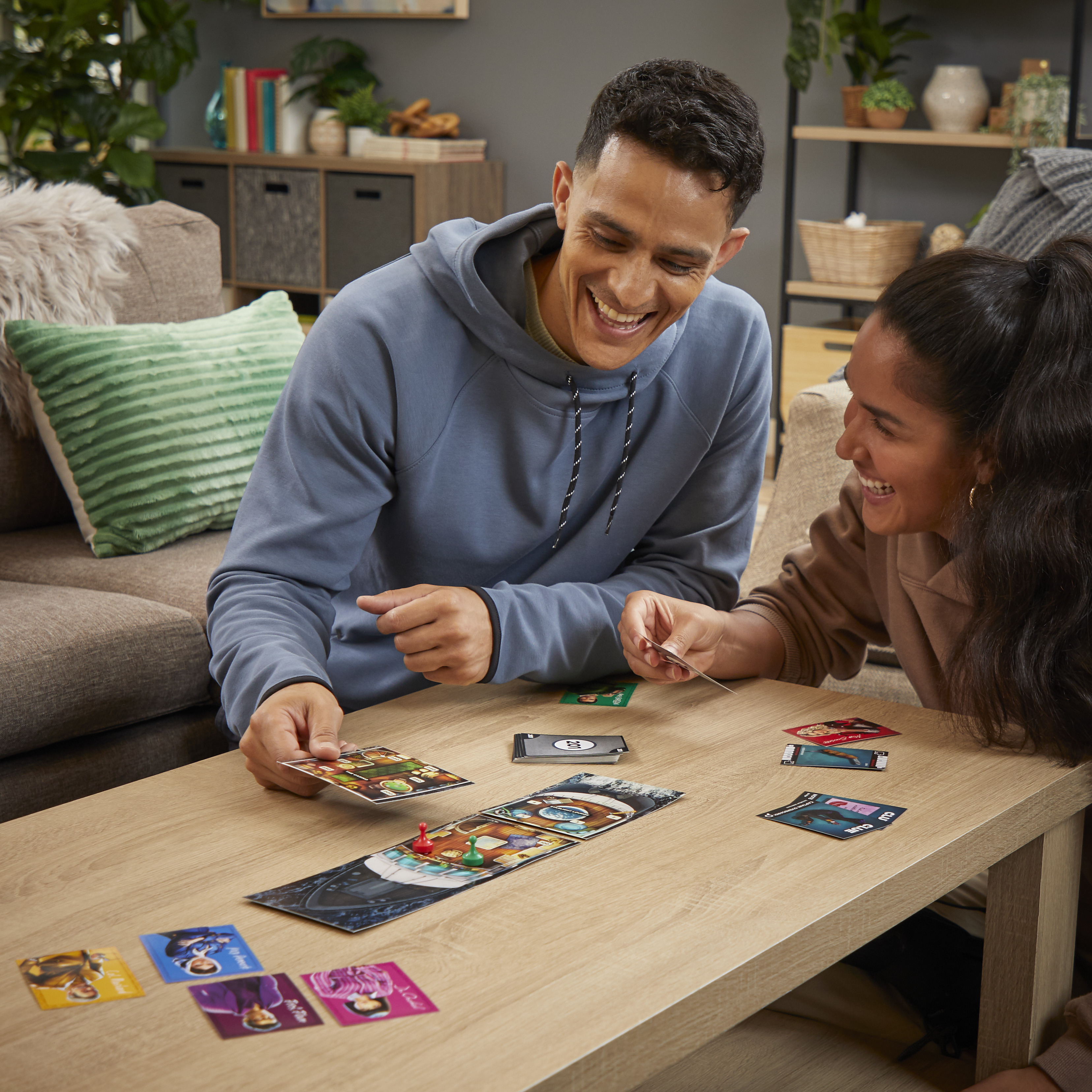 Zwei Personen spielen ein Brettspiel, lächeln. Karten und Spielbrett auf einem Holztisch.
