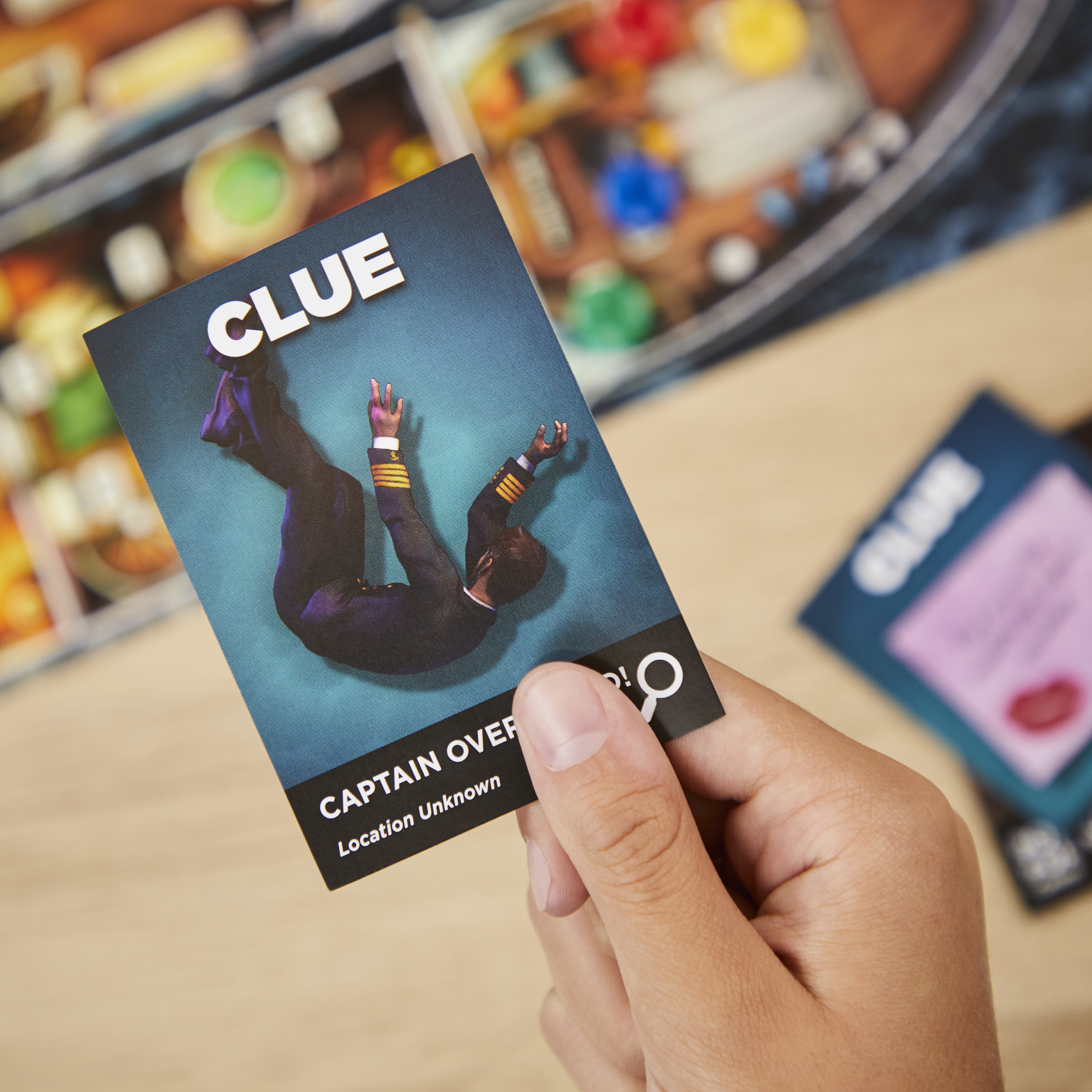 Person hält eine 'Cluedo'-Karte mit einer stürzenden Person darauf. Hintergrund Holztisch.