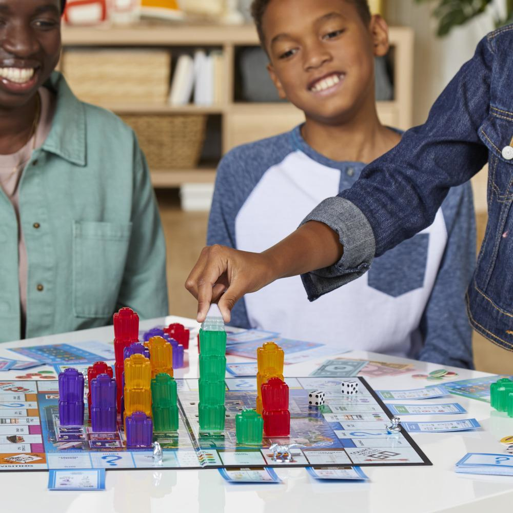 Drei Kinder spielen ein Brettspiel. Ein Kind platziert ein Gebäudeteil auf dem Spielbrett.