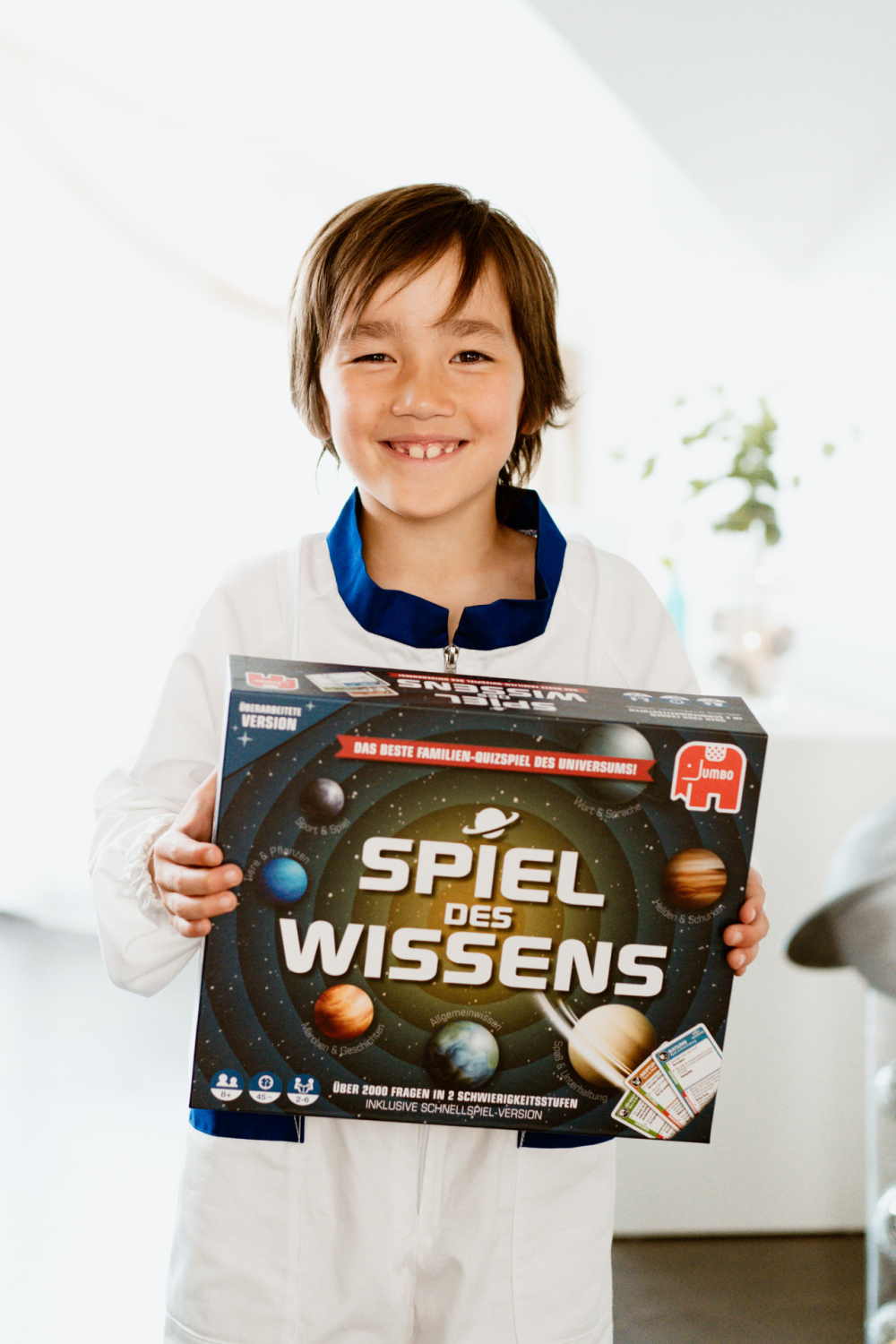 Junge in weißem Astronautenanzug hält ein Brettspiel mit Planetenbildern und deutschem Text.