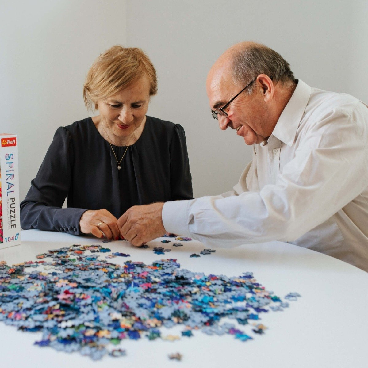 Dwie starsze osoby układają puzzle przy białym stole. Puzzle są na stole.