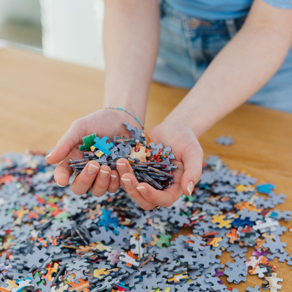 Hände halten einen Stapel Puzzleteile in verschiedenen Farben auf einem Holztisch, viele Teile verstreut.