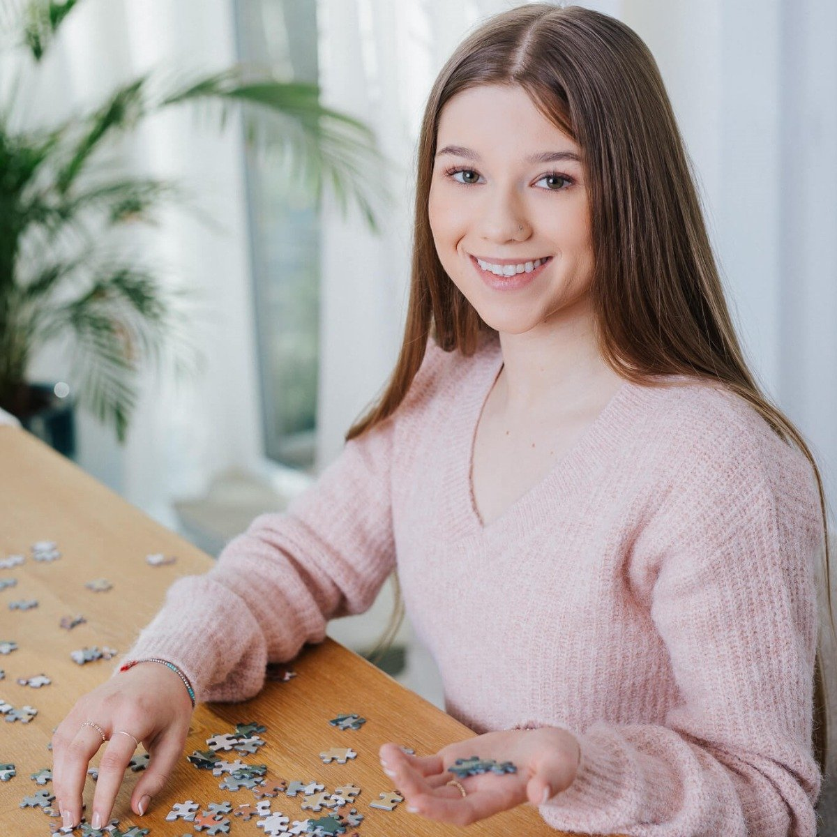 Eine junge Frau lächelt, während sie an einem Puzzle arbeitet, mit Teilen, die auf einem Holztisch verstreut sind.