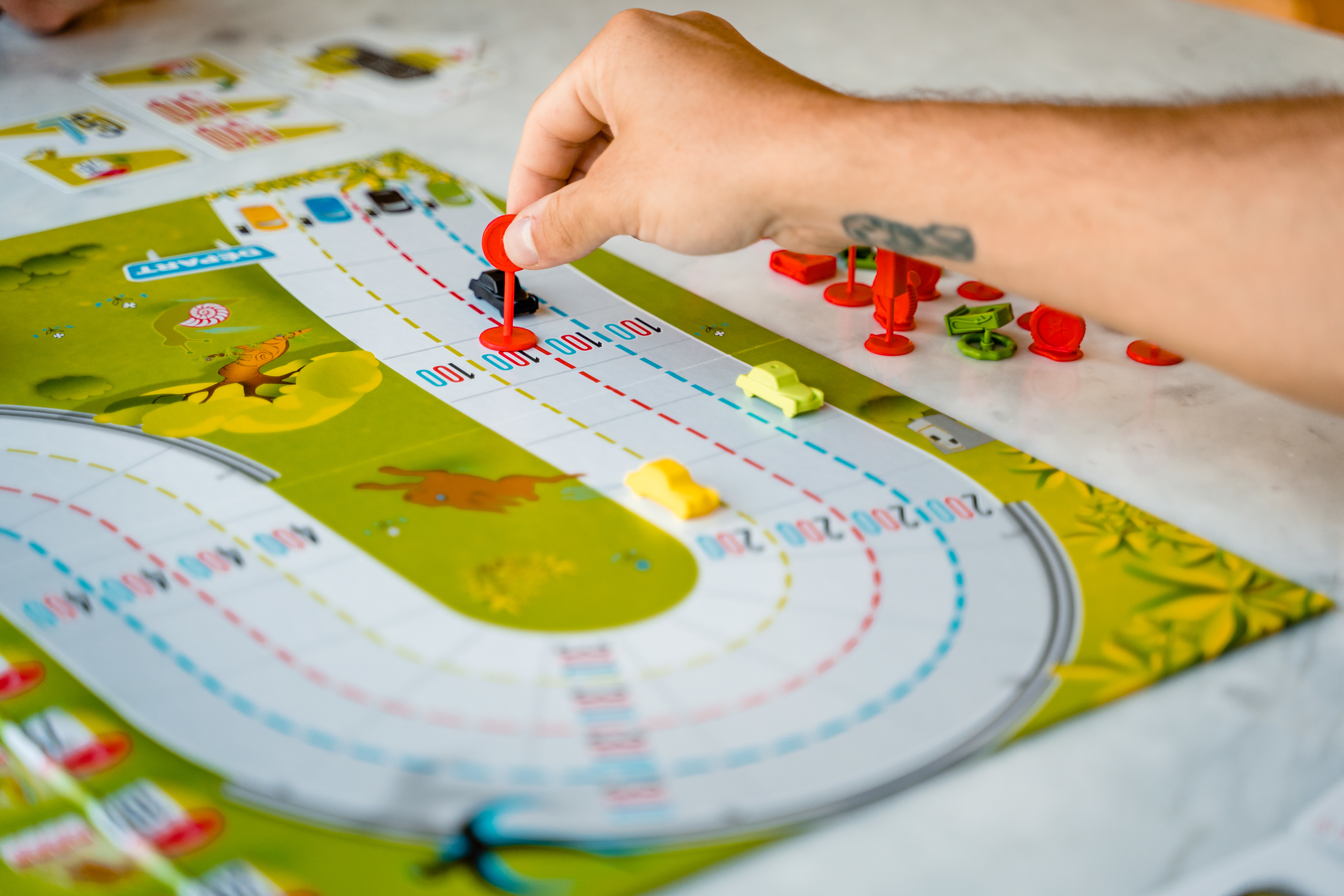 A person's hand moves a red car piece on a green board game with other cars.