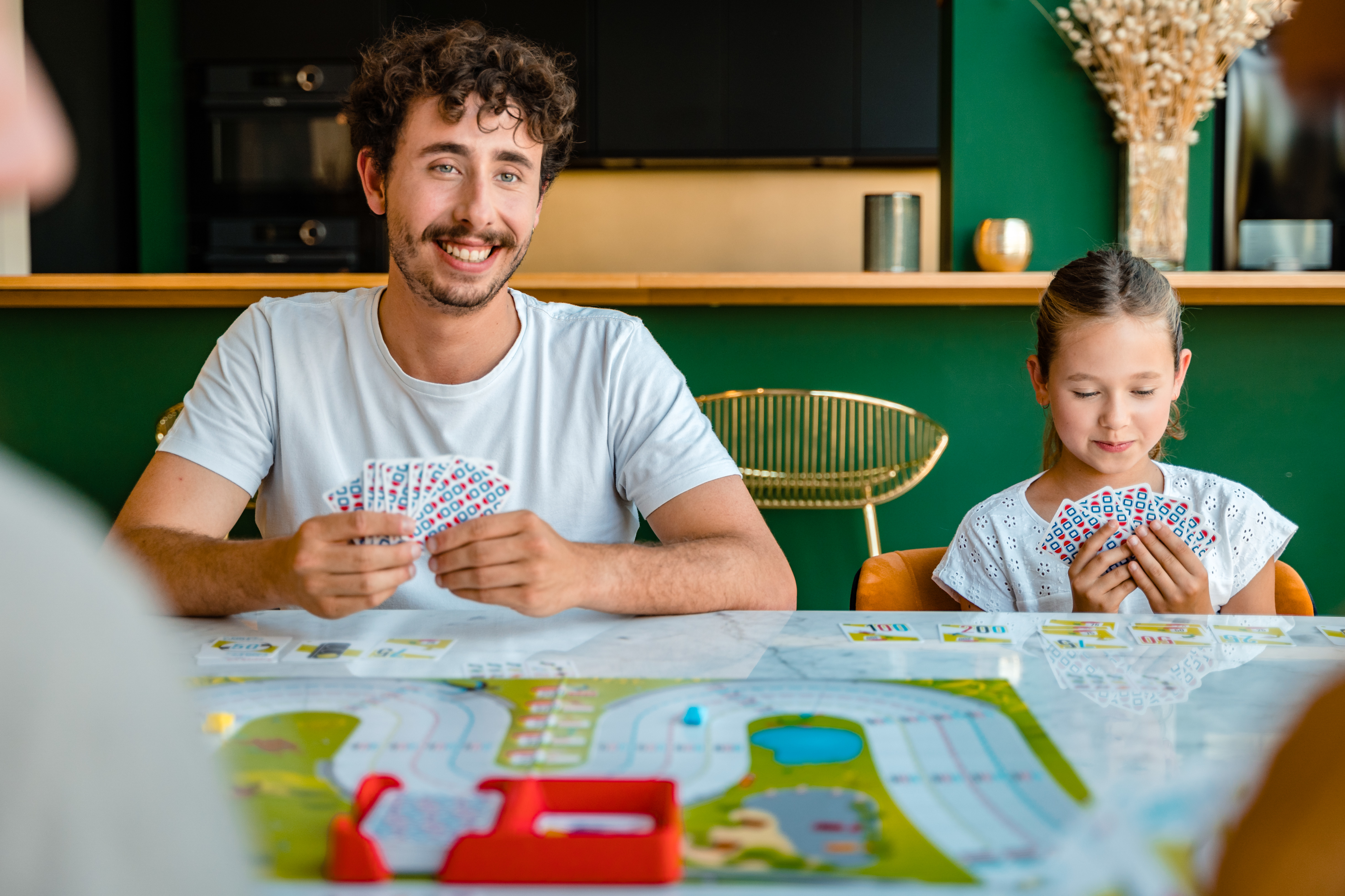 A man and a girl play cards at a table. Smiling man holds cards. Green wall in the background.